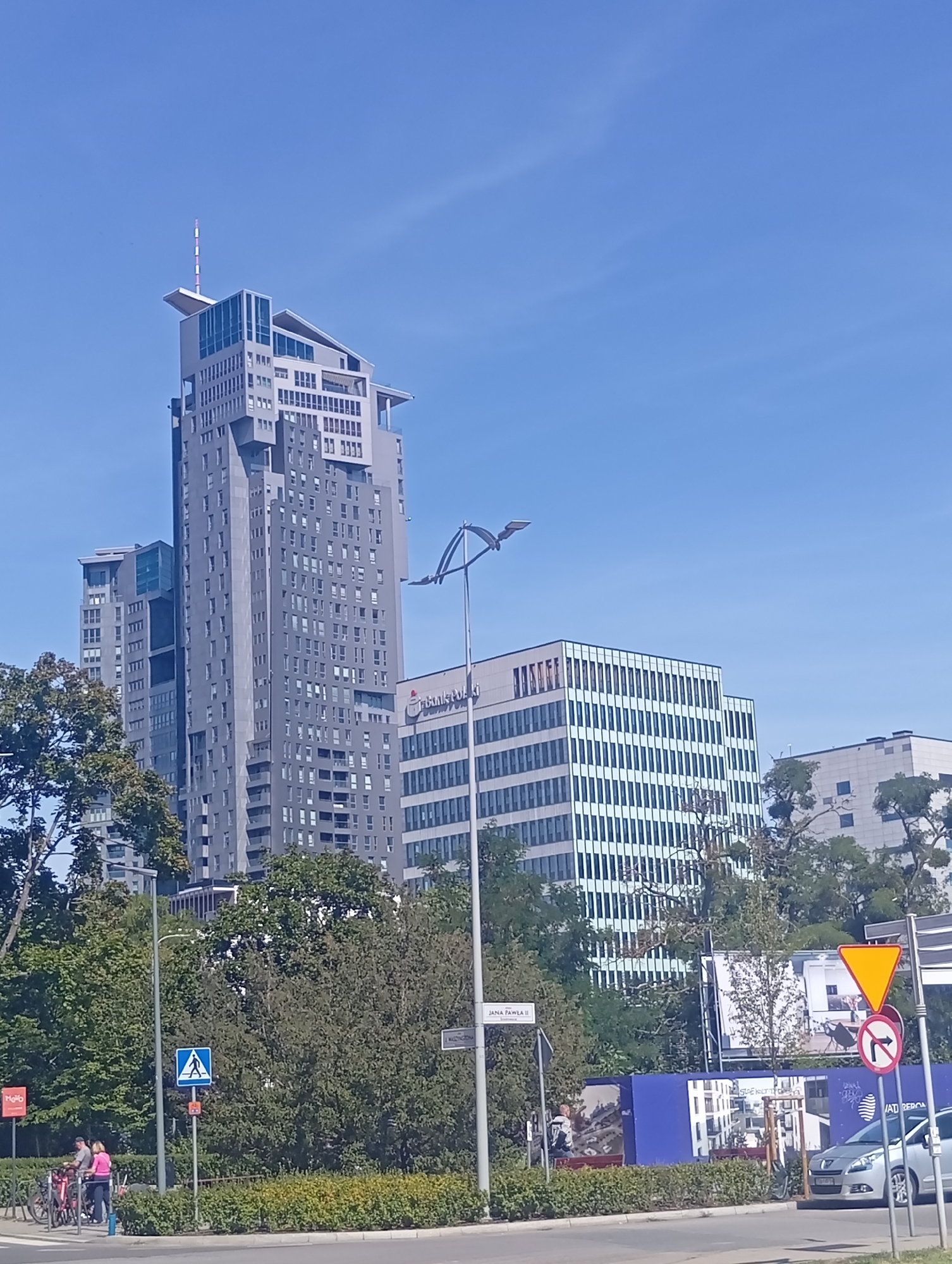 The image shows a view of the city of Gdynia, Poland, featuring the Sky Tower skyscraper in the background. The skyscraper is a tall, modern building with a distinctive design, featuring multiple levels and a stepped profile. In front of the skyscraper, there is a lower, rectangular building with a glass facade. To the left of the skyscraper, another tall building is partially visible.
The foreground of the image shows a street with a few people standing near bicycles and a parked car. There are also trees and bushes lining the street, as well as various street signs and traffic signals. The sky is clear and blue, indicating a sunny day.
Specifically, the Sky Tower is a prominent landmark in Gdynia, known for its height and unique architecture. The surrounding buildings appear to be a mix of commercial and residential properties. The presence of bicycles and people suggests that this is an urban area with active public spaces.
The overall impression is of a vibrant city scene with modern architecture and a lively atmosphere. The image captures a typical day in Gdynia, showcasing its urban landscape and the everyday activities of its residents.