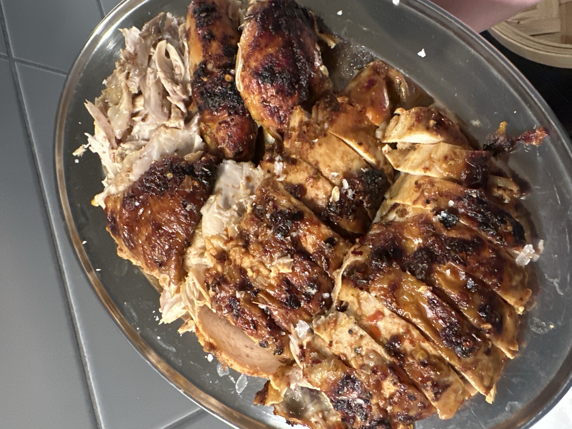 A silver serving platter is arranged with the remains of a roasted chicken. The chicken is partially shredded and sliced. There are light gray wall tiles visible in the background. The location is not discernible from the image.
