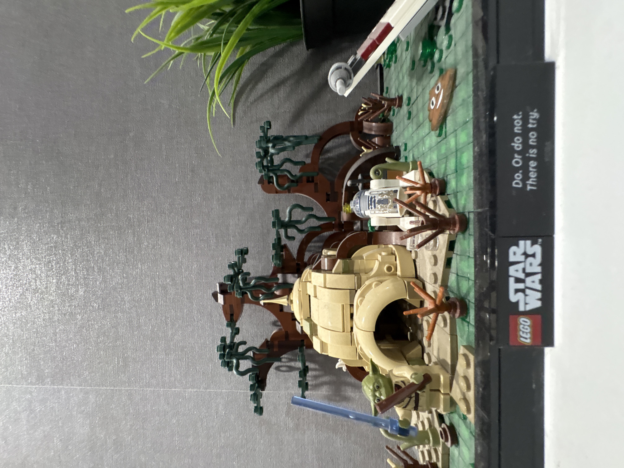A LEGO Star Wars Dagobah Diorama set, featuring Yoda's hut built within a tree and other swamp elements, is placed against a gray wall. To the upper left is green grass in a black planter, with the corner of a white table on the right side. The LEGO set includes a small X-wing fighter sunk in the swamp and a display plaque with the "LEGO Star Wars" logo and the quote "Do. Or do not. There is no try.". Yoda is represented as a minifigure near the base of the tree.