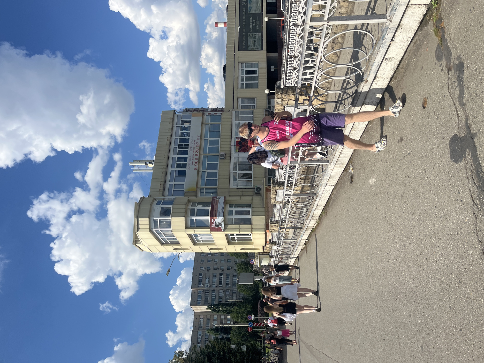 The image captures a sunny day in an urban setting, likely Krasnodar, Russia. A group of people are standing on a sidewalk next to a decorative white metal railing. The foreground features a person wearing a pink tank top with text "ФВКК" and "CH.VOLLEY.RU" visible, along with purple shorts and patterned clogs. They are posing with their hand on their hip and appear to be relaxed or perhaps posing for a photo. Behind them, more people are standing in a line, some of whom are women in dresses.
The background shows a multi-story building with many windows, some of which have air conditioning units. Another, older apartment building with rows of balconies is visible further in the distance, suggesting a typical Russian urban landscape. The sky is bright blue with fluffy white clouds, indicating clear and pleasant weather, and the strong shadows suggest it is midday. The overall scene appears to be a casual gathering or a group outing on a warm day.
Visible text includes "АРЕНДА ПРОДАЖ" (Rent/Sale), a phone number "8 989 828 97 07", and other Russian text related to a banquet hall and contact information on signs attached to the building. The scene conveys a sense of everyday urban life under a bright sky.