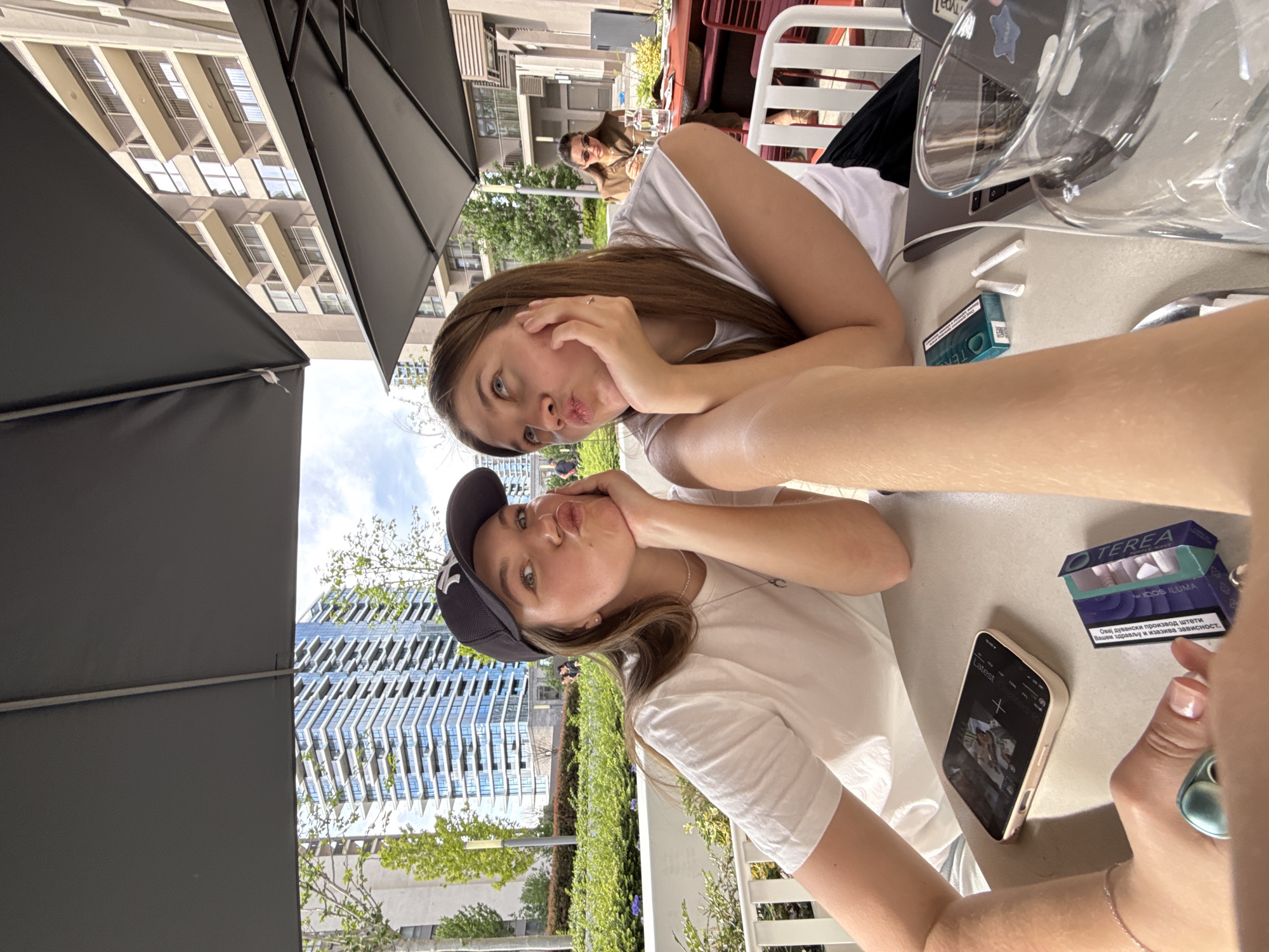Two young women pose cheek to cheek at an outdoor cafe table. The woman on the left wears a white t-shirt and black baseball cap. The woman on the right wears a white t-shirt. Both girls are making a kissing face. On the table is an iPhone, a pack of cigarettes (TEREA, IQOS ILUMA), cigarettes, a laptop computer, a glass of water, and an umbrella. Other people are visible in the background. The buildings indicate the location as Belgrade, Serbia.