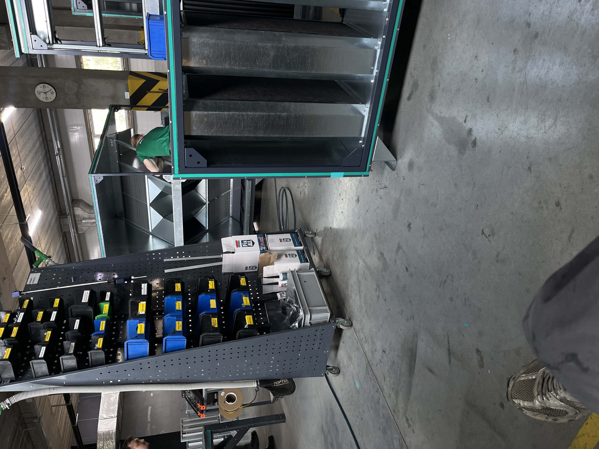 The image depicts a man wearing a green t-shirt working inside a large, industrial-style building, possibly a factory or warehouse. He is positioned near a tall, multi-tiered metal shelving unit that appears to be part of a ventilation system or a storage rack. The shelving unit has several compartments, some of which are filled with metallic components, while others are empty. To the left of the shelving unit, there is a circular clock on a concrete pillar. The overall scene suggests an indoor industrial environment.
In the foreground, a dark gray rolling cart is visible, laden with various items, including what appear to be tools and containers. Some of these items are blue, black, and yellow, with some having yellow labels attached. Boxes, some with "FIX ALL" printed on them, are also present on the cart. The floor is concrete, showing some wear and tear, with a few dark stains. A person's legs and feet, wearing athletic shoes, are visible in the lower right corner, indicating another person is present but mostly out of frame. The image is oriented vertically, suggesting it might have been taken with a phone camera. The lighting appears to be artificial, typical of an indoor industrial setting. There are no clear indicators of the time of day or weather conditions, but the indoor setting implies it is not directly affected by external weather. The man's posture suggests he is actively engaged in work or inspection related to the shelving unit.