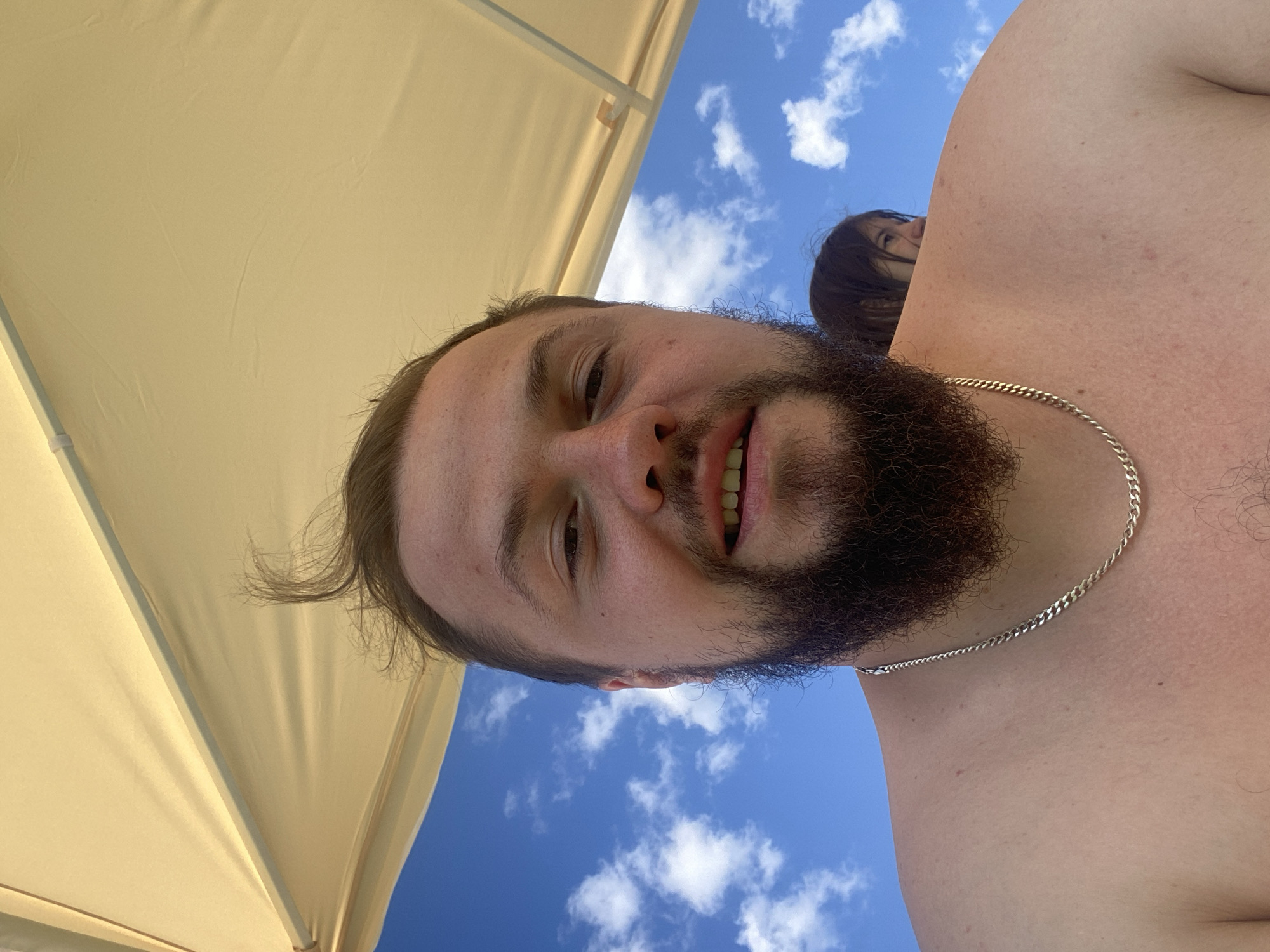 The image is a selfie taken outdoors under a tan umbrella. A man with a beard and wearing a silver chain is the main subject, smiling at the camera. Behind him, a person with dark hair can be seen, partially obscured by the man's shoulder. The background consists of a bright blue sky with scattered white clouds. The location is not specified in the image; it is not possible to determine if it is in Колокша, Russia.