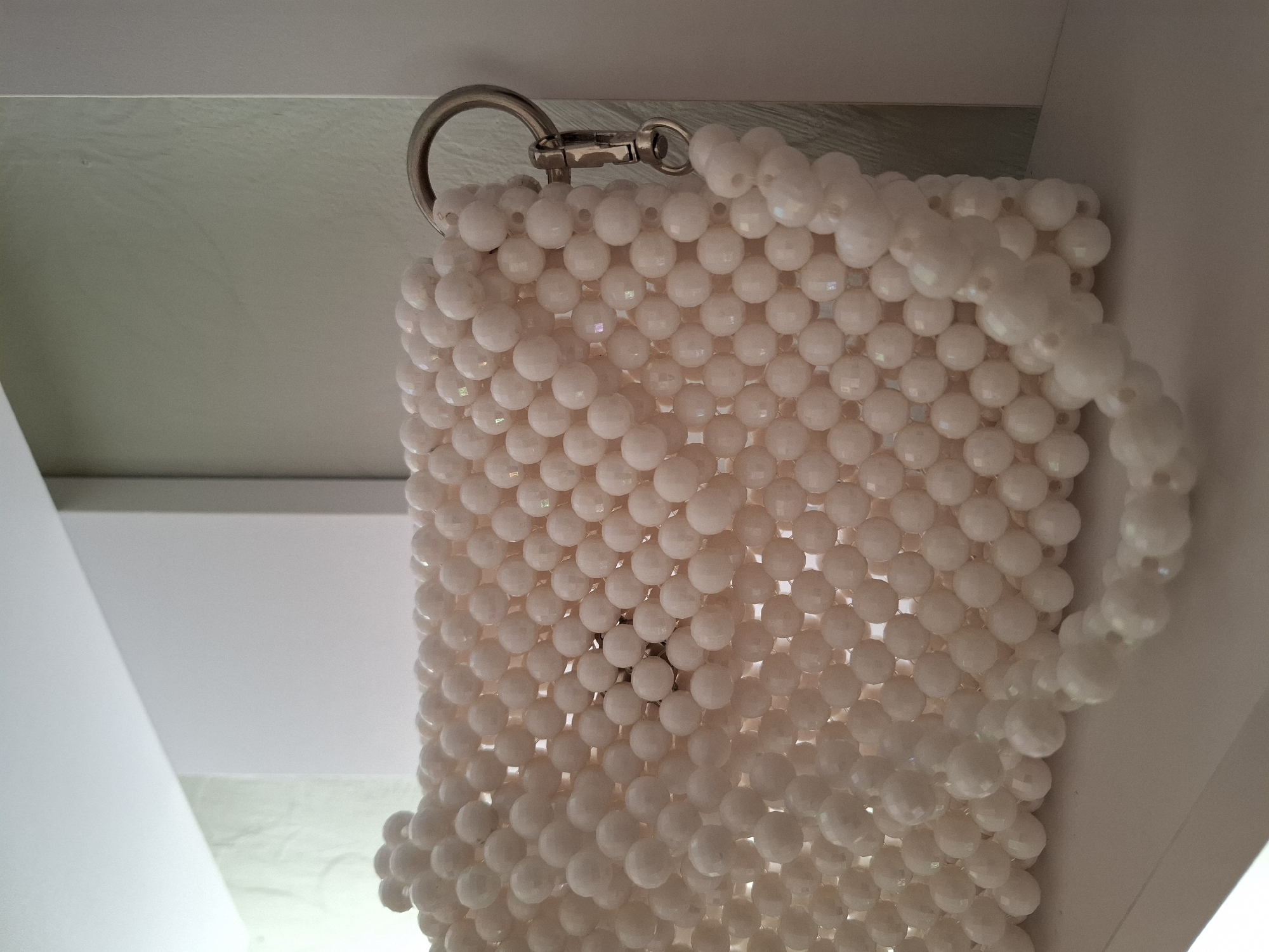 The image shows a close-up of a white beaded bag. The bag is made of many small, round, faceted white beads, giving it a textured and slightly iridescent appearance. A silver metal ring and clasp are attached to the top of the bag, suggesting it has a handle or strap. The bag is hanging, as indicated by the downward orientation of the bead strands.
The background is a neutral, light-colored surface, possibly a wall or ceiling, with some angled lines suggesting architectural elements. The lighting is soft and diffuse, creating subtle highlights on the beads. There is no indication of people or a specific activity taking place. The overall impression is one of a still life focused on the texture and detail of the beaded accessory. The location context provided, Korenovsk, Russia, does not offer any visual cues within the image itself. No text is visible.