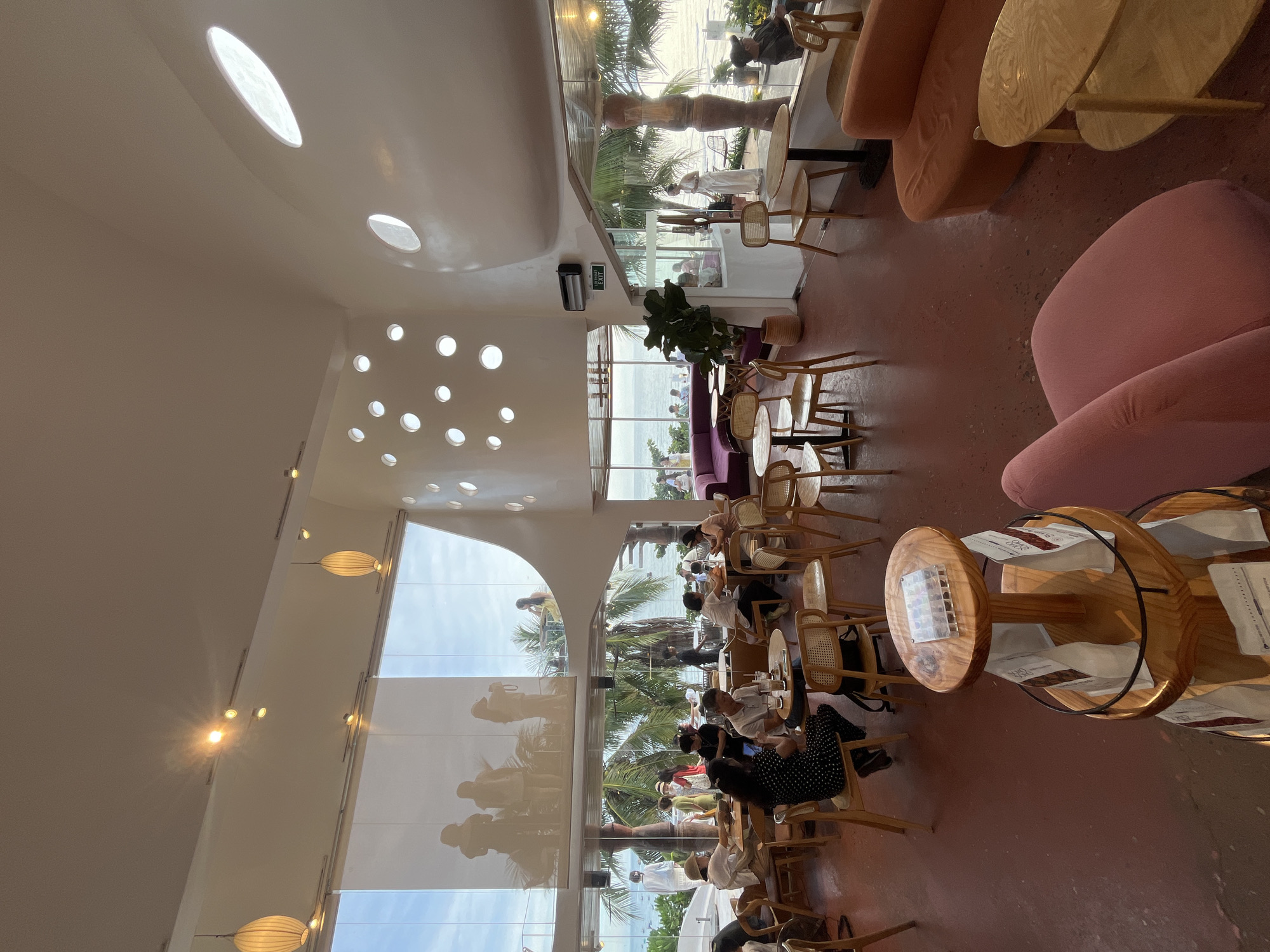 The image depicts the interior of a modern, brightly lit cafe situated in Mũi Né, Vietnam, with a prominent view of the ocean. The cafe features a curved, white ceiling with scattered circular cutouts that allow natural light to filter through. Large windows offer a panoramic vista of the sea, palm trees, and a distant shoreline with boats.
Inside, the cafe is furnished with a mix of seating arrangements. Several tables with light-colored wooden chairs, some with cane backs, are scattered across the reddish-brown speckled floor. In the foreground, a tiered wooden display stand holds various packaged goods, likely coffee or tea. Towards the back, a long, curved purple sofa faces the ocean view, and a few other tables and chairs are visible. Orange and pink upholstered sofas and armchairs are also present, adding pops of color to the space.
Numerous patrons are seated at tables, engaged in conversation or enjoying their beverages. Some individuals are standing, and others are walking through the cafe. On the upper level, visible through a large window, are silhouettes of people, possibly a group posing for a photo. The overall atmosphere appears relaxed and social. An "EXIT" sign is visible above a doorway in the background.