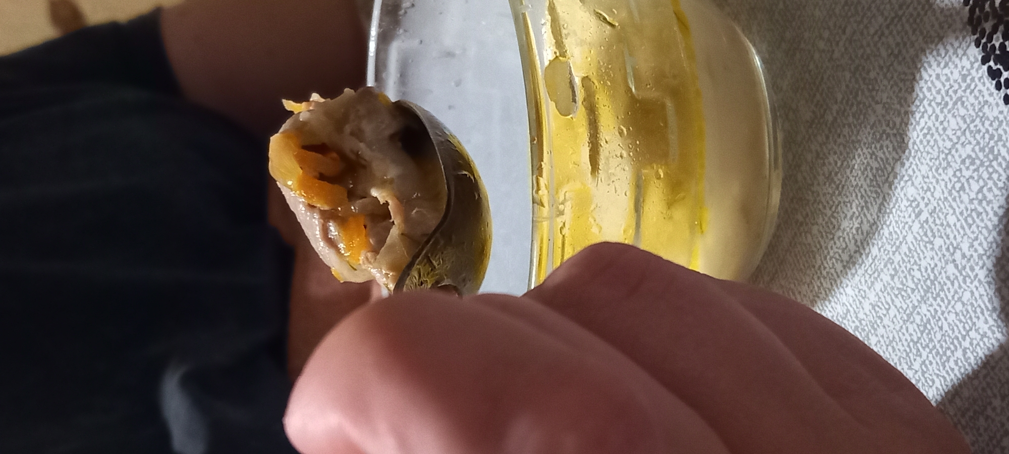 A close-up, slightly rotated indoor shot captures a person's hand holding a spoon filled with food. The food on the spoon appears to be a light-colored, possibly dumpling-like item with visible orange or yellow pieces, likely cooked vegetables such as carrots or squash. The metal spoon is held by a hand with visible knuckles and skin texture.
To the right of the spoon, a clear glass or plastic container is visible, featuring streaks and splashes of a thick yellow substance on its interior surface, which could be a sauce, puree, or a drink. Beyond the container, a patterned textile in shades of white, grey, and black is visible, suggesting a tablecloth or blanket. In the blurred background to the left, dark, indistinct shapes hint at clothing or furniture. The lighting is somewhat dim, indicative of an indoor setting, possibly with artificial light. No specific location cues are present within the image itself that relate to Kryvyi Rih, Ukraine, beyond the provided context. There is no visible text. The scene depicts the act of eating.