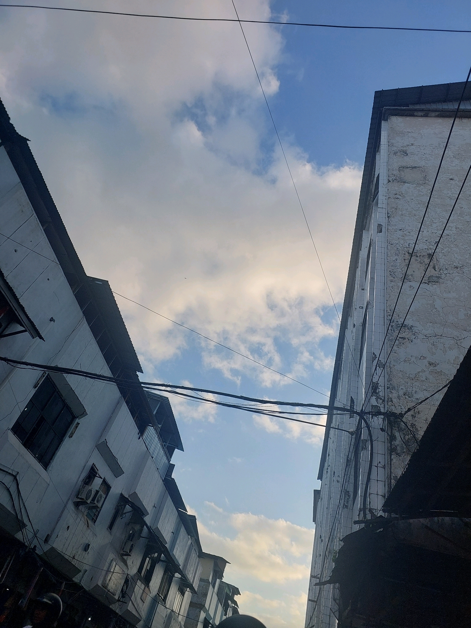 The image is a low-angle shot looking up at buildings and the sky in Ambon, Indonesia. Two buildings, one on the left and one on the right, dominate the frame, creating a narrow alleyway perspective. The building on the left appears to be a residential or commercial structure with multiple floors, visible windows, and what looks like air conditioning units. Its exterior is a light color, and it has a corrugated metal roof. The building on the right is taller and seems to be made of concrete, with a textured, somewhat aged facade. It also has a corrugated roof. A dense network of electrical wires crisscrosses the sky between the buildings. The sky is partly cloudy with patches of blue and white clouds, suggesting daytime. The overall impression is of a dense urban environment, possibly an older part of the city, with visible signs of wear and tear on the buildings and a utilitarian arrangement of overhead wiring. There are no people clearly visible in the frame, but the presence of the buildings and infrastructure suggests human activity.