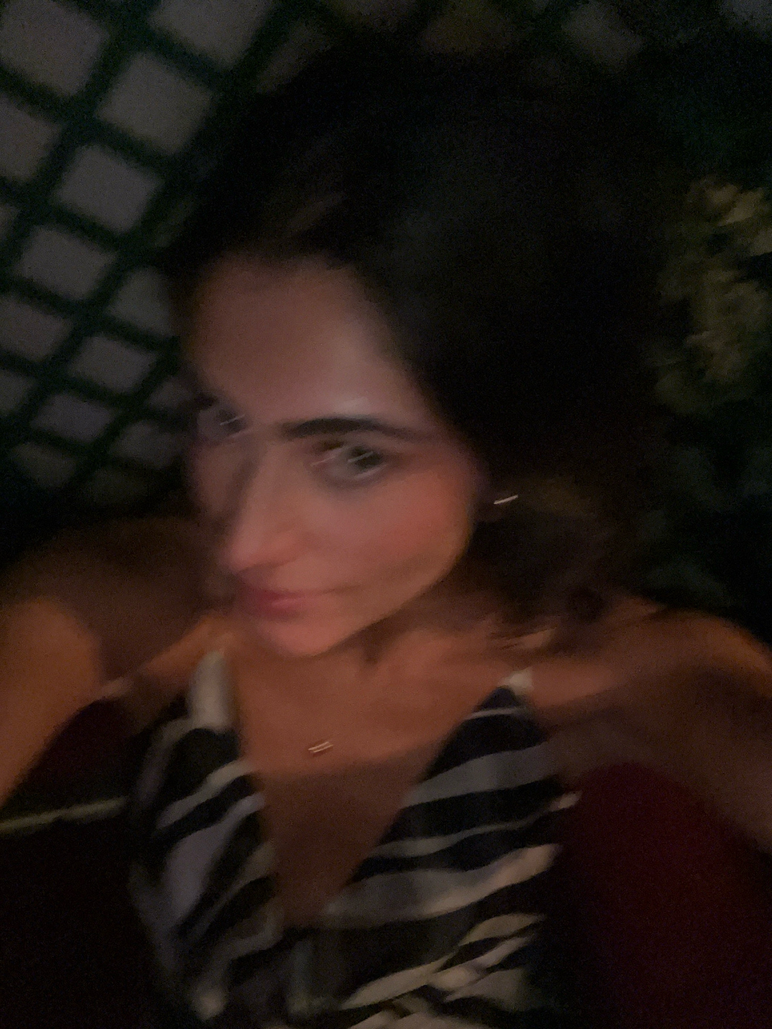 The image is a close-up, blurred selfie of a woman. She has dark hair and is wearing a patterned top. She appears to be indoors, possibly in a dimly lit room with a lattice-like structure visible in the background. There is no indication of the city of Madrid, Spain, or any specific location.