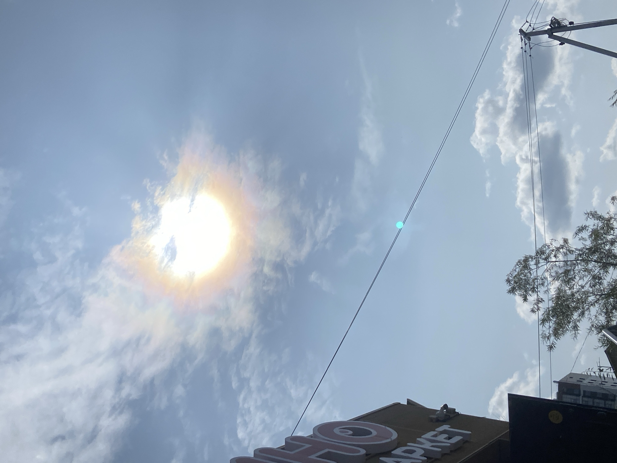 The image shows a bright, sunny sky with scattered clouds. The sun is visible, with a halo-like effect around it, suggesting the presence of ice crystals in the atmosphere, possibly indicating a sun dog. In the foreground, part of a building with signage is visible on the left, with the letters "HO" and "AP" of a larger word being partially obscured. Two power lines stretch horizontally across the frame. A small, bright turquoise object is visible on the upper power line. In the lower part of the image, a tree with sparse foliage is seen, along with a distant building and a utility pole. The overall scene suggests an outdoor urban environment under a bright sky, consistent with a location in Yakutsk, Russia, during daylight hours.