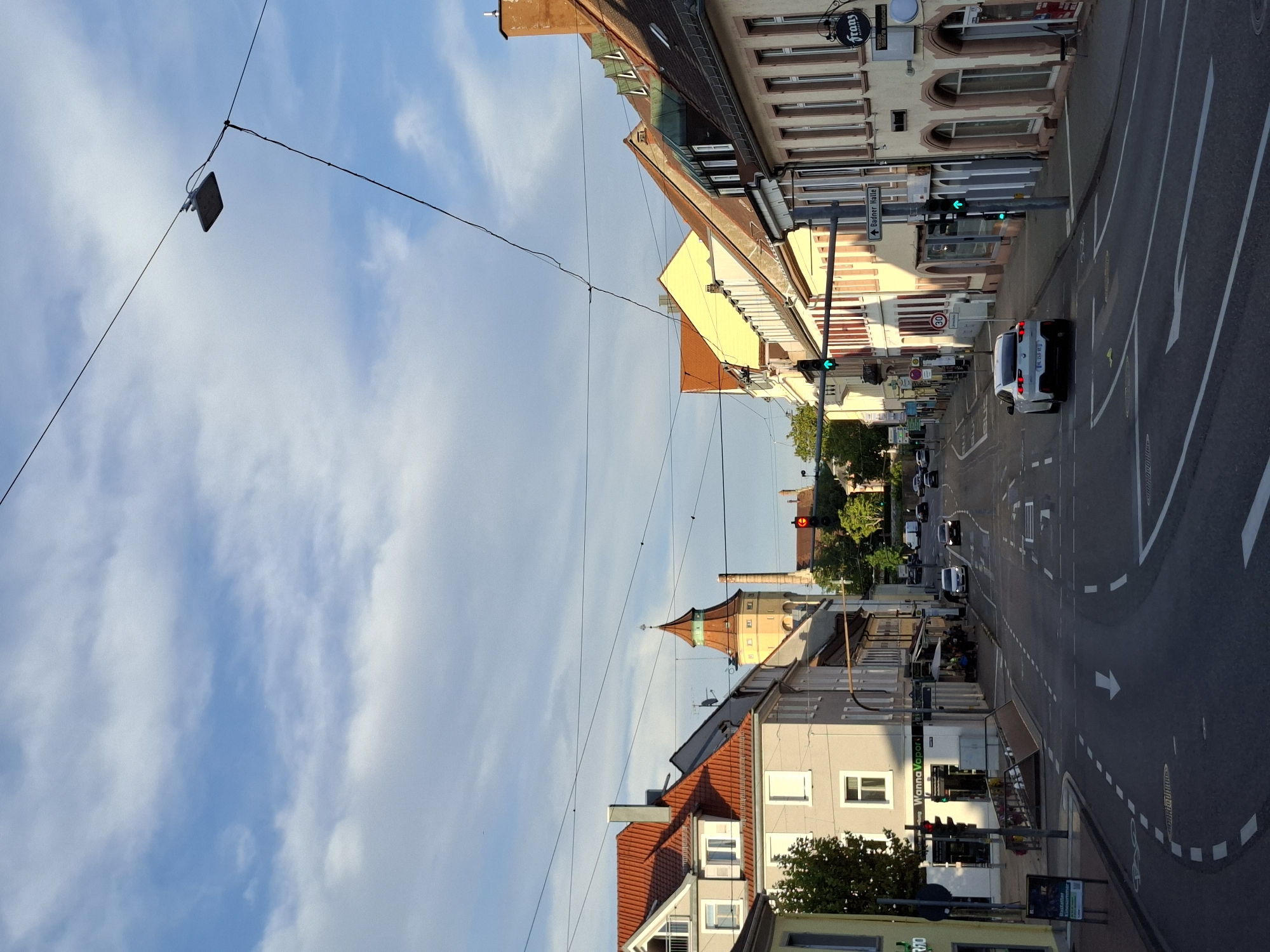 The image depicts a street scene in Rastatt, Germany, likely during daylight hours with a partially cloudy sky. The perspective is from a slightly elevated position, looking down a street lined with buildings. On the left side of the street, there are multi-story buildings with red tiled roofs and pale facades. A sign on one building reads "Wanna Vapor". Further down the street, a distinctive tower with a red conical roof is visible, along with a brick chimney. On the right side of the street, there are more buildings with tiled roofs and cream-colored facades, featuring arched windows. A sign on one of these buildings says "Franz".

The street is paved and has lane markings indicating traffic flow. Several cars are present on the road, including a white hatchback in the foreground. Traffic lights are visible, with some showing green signals. There are also power lines and a street lamp suspended across the street. The overall environment appears to be an urban or town center with commercial establishments and residential buildings.