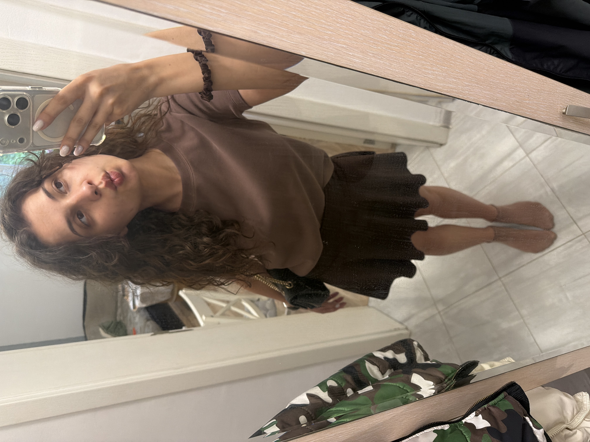 A young woman with curly brown hair is taking a mirror selfie. She is wearing a brown t-shirt and a brown pleated skirt. She has a dark scrunchie on her left wrist. She is holding a smartphone in her right hand. Her lips are puckered. The background appears to be a room with tiled flooring. A camouflage jacket is visible in the lower part of the image. The lighting suggests it could be daytime.