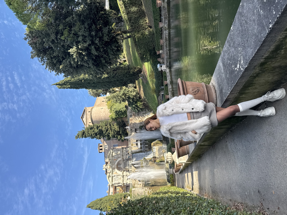 A female subject is depicted standing in the gardens of Villa d'Este in Tivoli, Italy, under a clear blue sky with sparse clouds. The subject, appearing to be of East Asian descent, stands on a paved pathway, leaning her right hip against a low, grey stone ledge that borders a calm, greenish reflecting pool on the right.
The subject wears a white, textured, collared faux fur jacket over a white, ribbed crop-top with a high neckline. Below, she wears a black and white plaid mini-skirt, sheer hosiery, white knee-high socks, and white platform sneakers with thick soles. Her long, light brown hair is visible, and her head is turned to her right, away from the camera. Her hands are in her jacket pockets.
In the mid-ground, to the left of the subject, a large, multi-tiered Baroque fountain is actively cascading water, flanked by tall cypress trees and manicured hedges. Further up the hill in the background, the historic Villa d'Este building, constructed of brick with a prominent rounded tower and other architectural elements, is visible amidst lush green foliage. The scene is illuminated by bright daylight, casting visible shadows.
