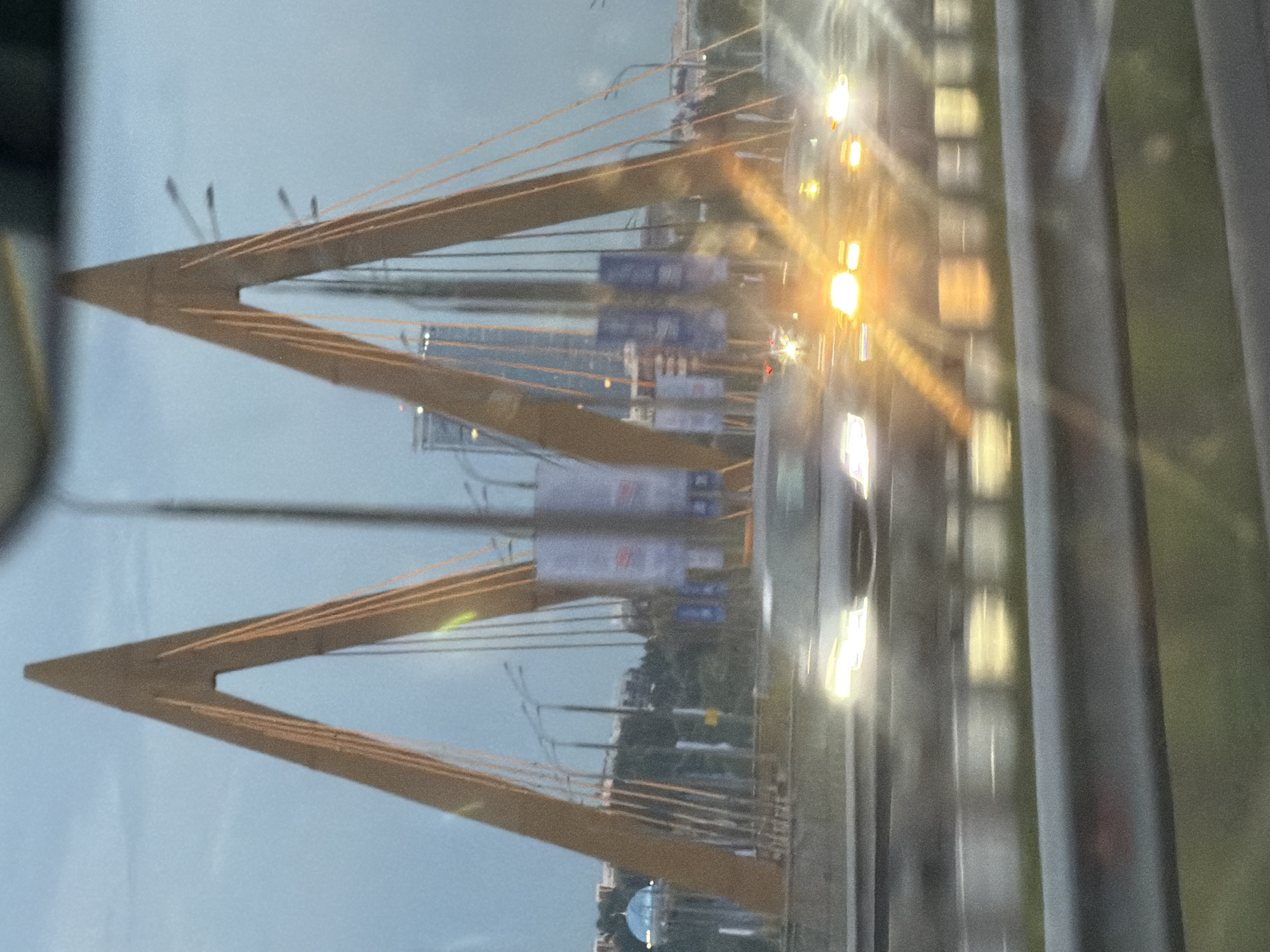 The image shows a view of the Millennium Bridge in Kazan, Russia. The bridge's distinctive yellow pylons and suspension cables are visible, spanning across a body of water. The scene is captured at dusk, with artificial lights from the bridge and surrounding city illuminating the darkening sky. Reflections of lights on the wet road surface suggest recent rain or ongoing precipitation.
In the background, the cityscape of Kazan is partially visible, with buildings and infrastructure hinting at an urban environment. The image appears to be taken from a moving vehicle, as indicated by the motion blur and the reflective surfaces that suggest looking through a window. The overall mood is somewhat melancholic due to the overcast sky and twilight setting, but the bright lights add a sense of vibrancy and life to the scene.
There are no identifiable people in the image. The primary objects are the bridge structure, cables, road, and distant city buildings. The weather appears to be overcast and possibly wet. The time of day is clearly evening or twilight. The location is confirmed as Kazan, Russia, and the Millennium Bridge is a recognizable landmark. No legible text is visible within the frame.