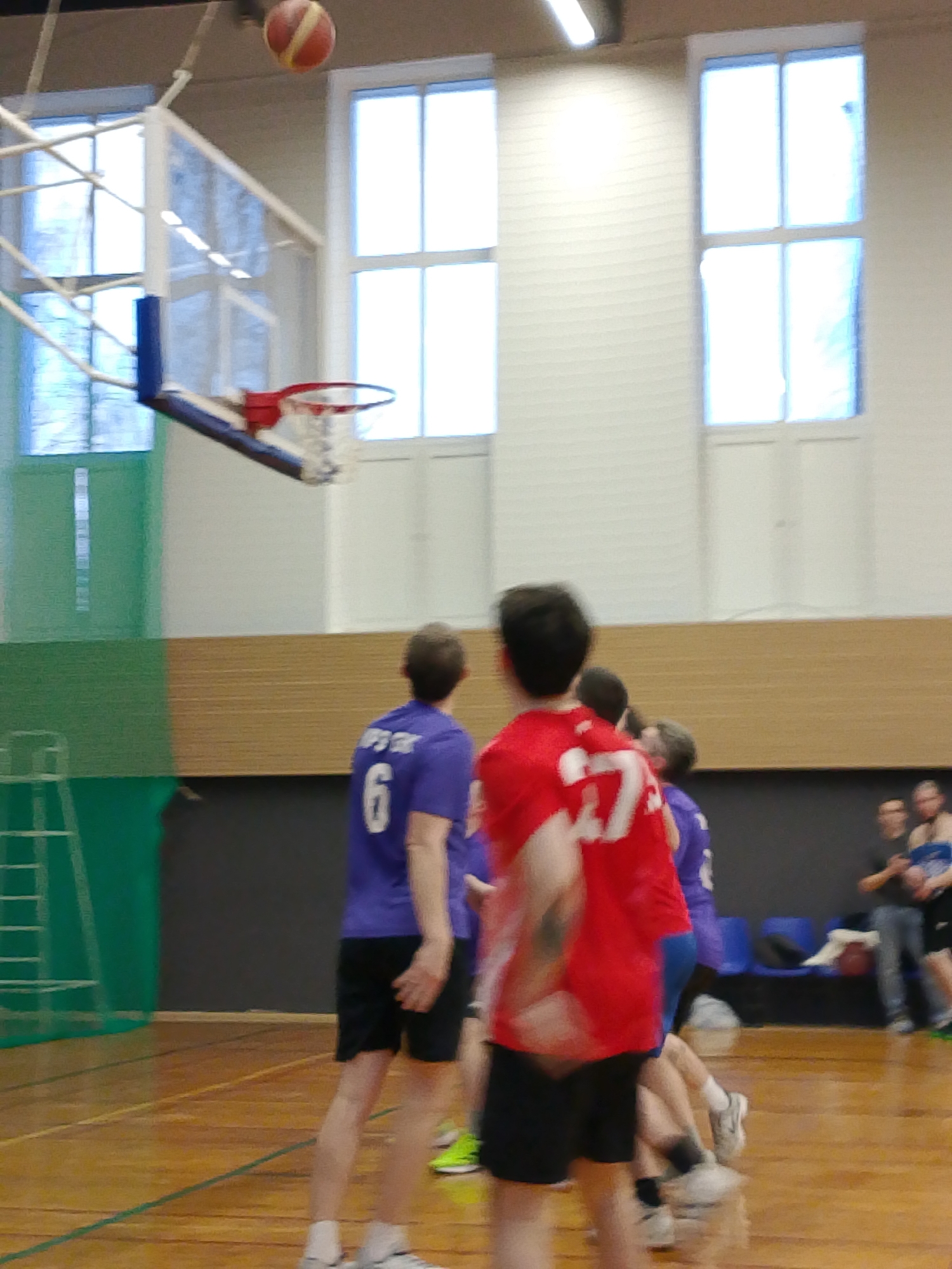 A basketball is in the air above a basketball hoop in an indoor gymnasium, likely during a game. The floor is wooden and has lines. Several players are visible, some in purple jerseys and one in a red jersey with the number 27. Spectators can be seen sitting in the background on blue plastic chairs. The gym has large windows and a wooden paneling along the walls. The location is Izhevsk, Russia.