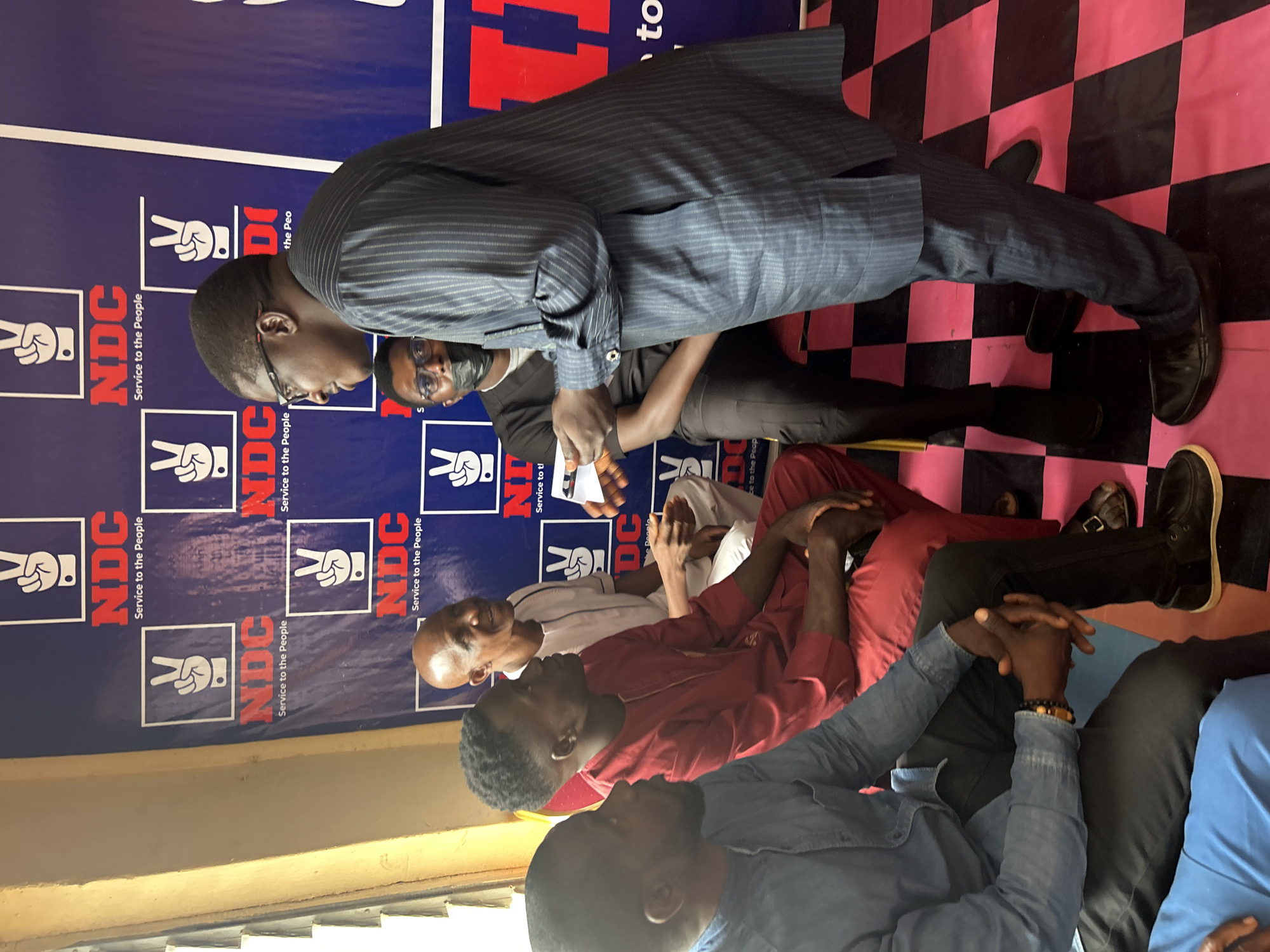 The image shows a group of people in what appears to be a meeting or gathering in Jos, Nigeria. One man, dressed in a pinstripe suit, stands and leans forward, seemingly addressing or interacting with the seated individuals. Behind them is a backdrop with the acronym "NDC" and the slogan "Service to the People" repeated, suggesting a political or organizational context. The floor has a pink and black checkered pattern. Some of the seated individuals are dressed in casual attire, and there is an interaction taking place between the standing man and at least one of the seated individuals. Some individuals appear to be in conversation or engaged in the meeting. The lighting in the room is bright, and there is a window visible in the background.