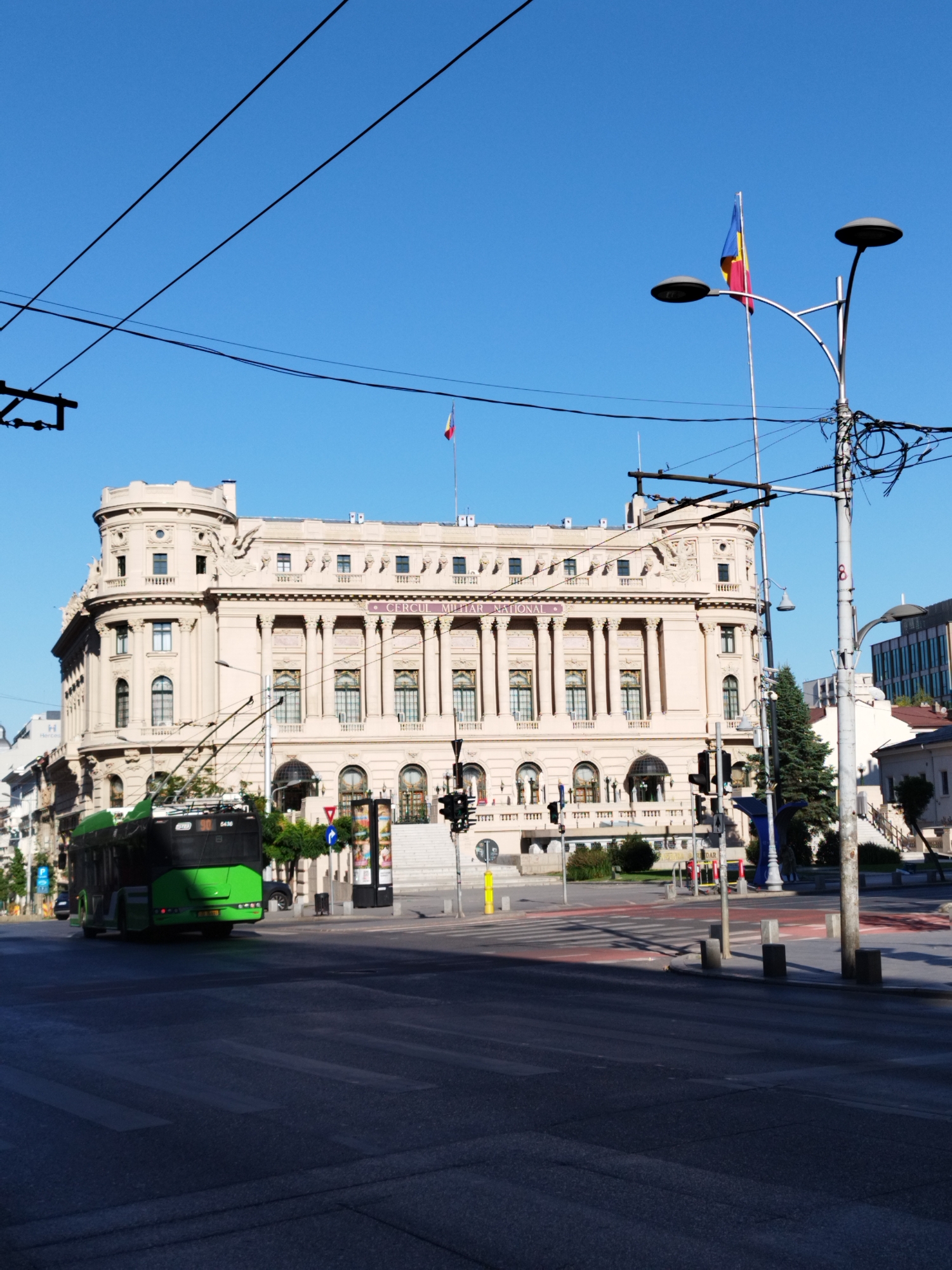 The image captures a view of the Cercle Militaire National in Bucharest, Romania, a grand neoclassical building with a prominent tower on the left side. The building is light beige with elaborate architectural details, including columns and ornate carvings. In the foreground, a green trolleybus, identified by the number "30" on its front, is moving to the left, crossing a street. The street features a pedestrian crossing and traffic lights. Overhead, a network of black wires crisscrosses the bright blue sky, indicating an urban setting with overhead electrical infrastructure. A Romanian flag waves from a flagpole atop a light post on the right side of the building. The scene appears to be captured in daylight, with shadows indicating the sun is positioned to the right of the viewer.