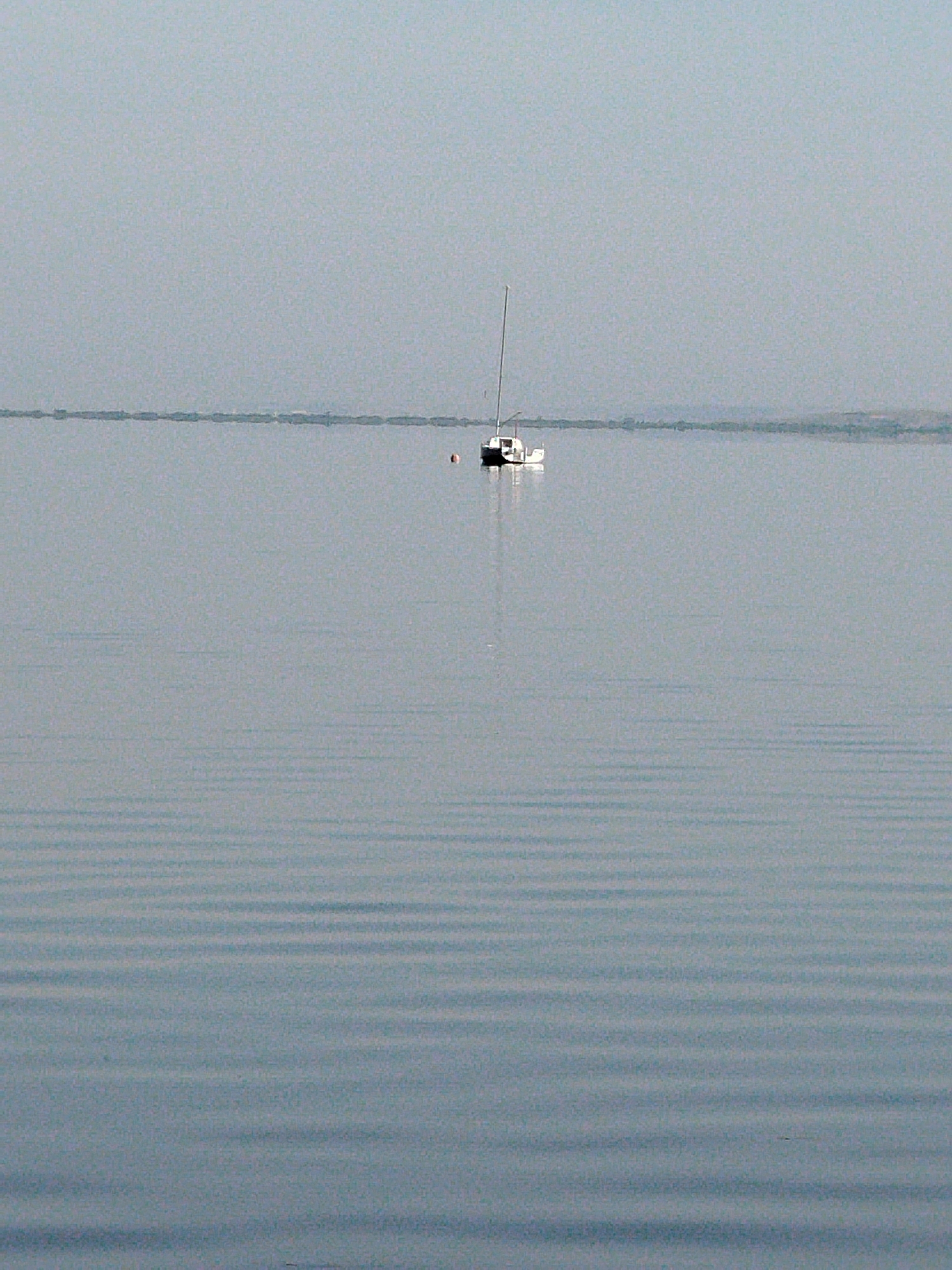 A sailboat with a single mast is moored on a calm, overcast body of water. The water exhibits subtle, horizontal ripples, creating a textured surface. The horizon is low and indistinct, with a faint, hazy outline of land visible in the distance. The sky is a uniform pale gray, devoid of clouds. A small, red buoy is visible near the sailboat. The overall scene conveys a sense of stillness and tranquility. The location is a large, natural body of water, possibly a lake or reservoir, consistent with the geography of Kokshetau, Kazakhstan, which is situated on the shores of Lake Kokshetau.