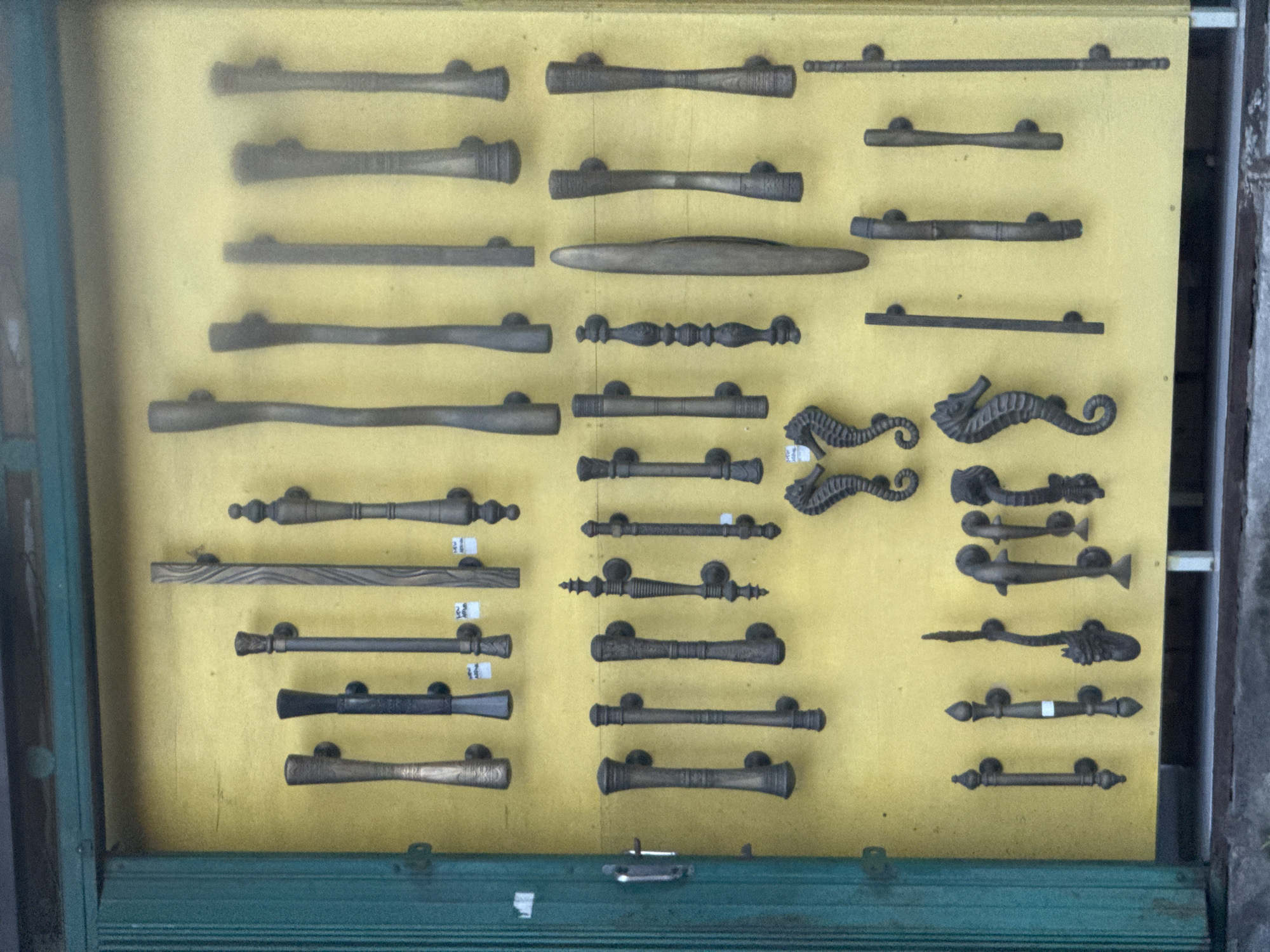 The image displays a collection of various antique-style metal door handles or pulls, meticulously arranged on a vibrant yellow display board. The handles appear to be made of dark metal, possibly aged bronze or cast iron, suggesting craftsmanship and durability. They are presented within what seems to be a display case, framed by a greenish-blue border at the bottom and left side, indicative of a shop or showroom setting.
The handles exhibit a wide range of designs, from simple, cylindrical bars to more elaborate and ornate shapes. Several styles feature intricate patterns, twisted motifs, or textured surfaces. Notable designs include a pair of handles shaped like seahorses, and another pair resembling stylized fish, showcasing marine-themed artistry. Other handles incorporate decorative knobs, ridged sections, and flared ends.
Small white rectangular labels or stickers are visible on some of the handles, likely indicating product codes, prices, or inventory numbers. For instance, a label with the number '9' is attached to one of the seahorse handles, while another on the left side shows '12'. A label with '24' is seen on a handle in the middle section, and '35' on a handle on the right. The overall impression is that these items are for sale or exhibition. The scene takes place in North Kuta, Indonesia, suggesting these may be local crafts or imports sold in the region.