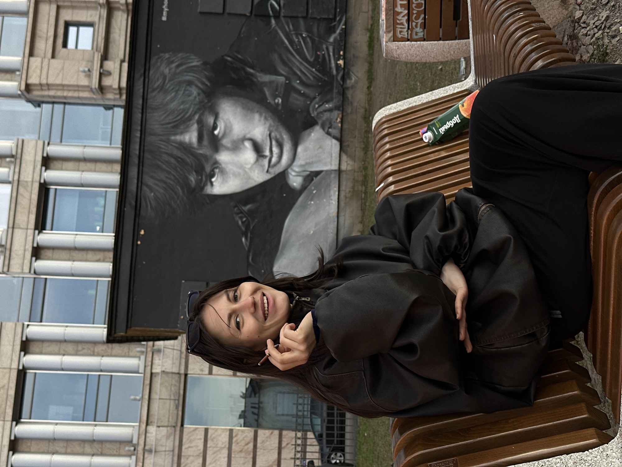 A young woman with long dark hair and sunglasses perched on her head is seated on a wooden bench in what appears to be Saint Petersburg, Russia. She is wearing a black leather jacket and black pants, smiling broadly at the viewer while holding a lit cigarette in her hand. Her left hand rests on her lap.
To her left, a large black and white mural of a man's face in a leather jacket is painted on a dark wall. Part of a hashtag, "#myha," is faintly visible above the mural. In the background, a modern building with multiple windows and light-colored architectural elements stands tall.
Next to the woman on the bench, a green "Добрый" (Dobryi, Russian for "Good") brand juice box, featuring an image of fruit (likely peach), is visible. On another bench in the upper right, partially obscured graffiti-like text "RU_NT" and "SSC.D" can be seen. The overall setting suggests a public urban space, possibly a park or square, during daytime, with a slightly overcast sky. The image is taken at a relaxed moment, capturing the woman's cheerful expression.