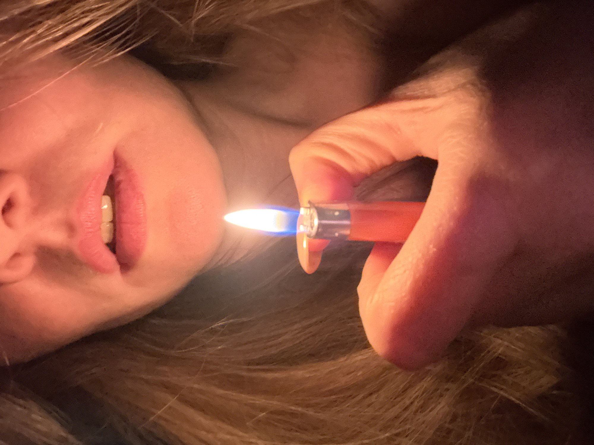 A close-up, inverted view shows a person's face and hand holding a lit orange lighter. The lighter's flame illuminates the person's face and hair. The image does not provide information to determine a specific location, such as Chaikovsky, Russia.