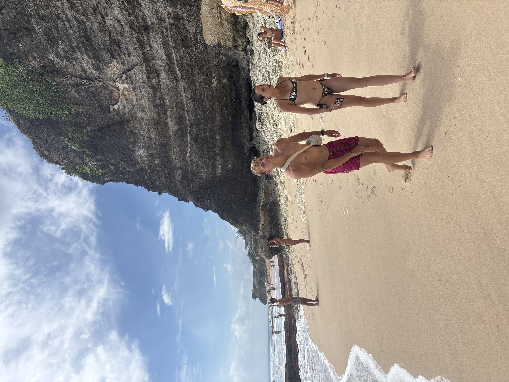 In Kuta Selatan, Indonesia, on a sandy beach with gentle waves lapping the shore, a couple walks along the wet sand. The man, wearing pink patterned shorts, walks to the left, and the woman, in a black and white polka dot bikini, walks to his right, looking forward. Behind them, a steep, rocky cliff face covered in green foliage rises majestically. Other beachgoers can be seen in the distance, some wading in the water, others relaxing on the sand. The sky is a clear blue with scattered white clouds. The overall scene depicts a sunny day at a popular tourist destination.