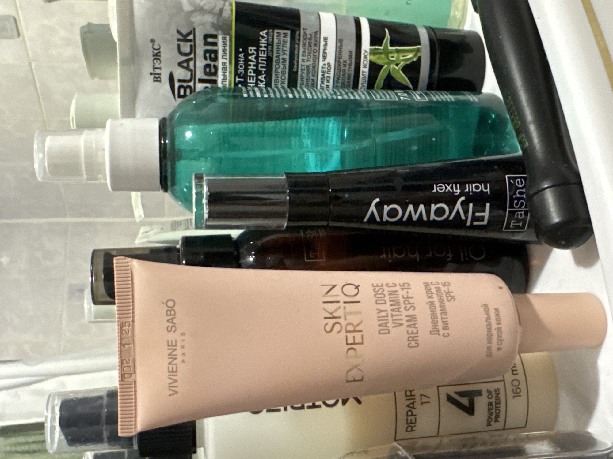 The image displays a close-up, eye-level view of various cosmetic and personal care products arranged on a shelf or surface, likely in a bathroom setting. The products are predominantly bottles and tubes with visible branding and text.
In the foreground, a light pink tube labeled "VIVIENNE SABÓ PARIS SKIN EXPERTIQ DAILY DOSE VITAMIN C CREAM SPF-15" is prominently featured. To its left, a beige bottle with the label "REPAIR 17 4 POWER OF PROTEINS 160 m." is partially visible. A black bottle with white text reading "Flyaway hair fixer Tashe" is to the right of the pink tube. Behind these, a tall, transparent green bottle with a white pump dispenser is visible, and to its right, a black and white tube labeled "ВІТЭКС BLACK clean" is present. Smaller bottles and containers are also scattered throughout the background, some with translucent caps. The overall lighting suggests an indoor environment, possibly with overhead lighting. The arrangement of the items implies a personal collection or display of beauty products. There is no indication of the specific location within Яромінскі сельскі Савет, Belarus, as the image focuses solely on the products.