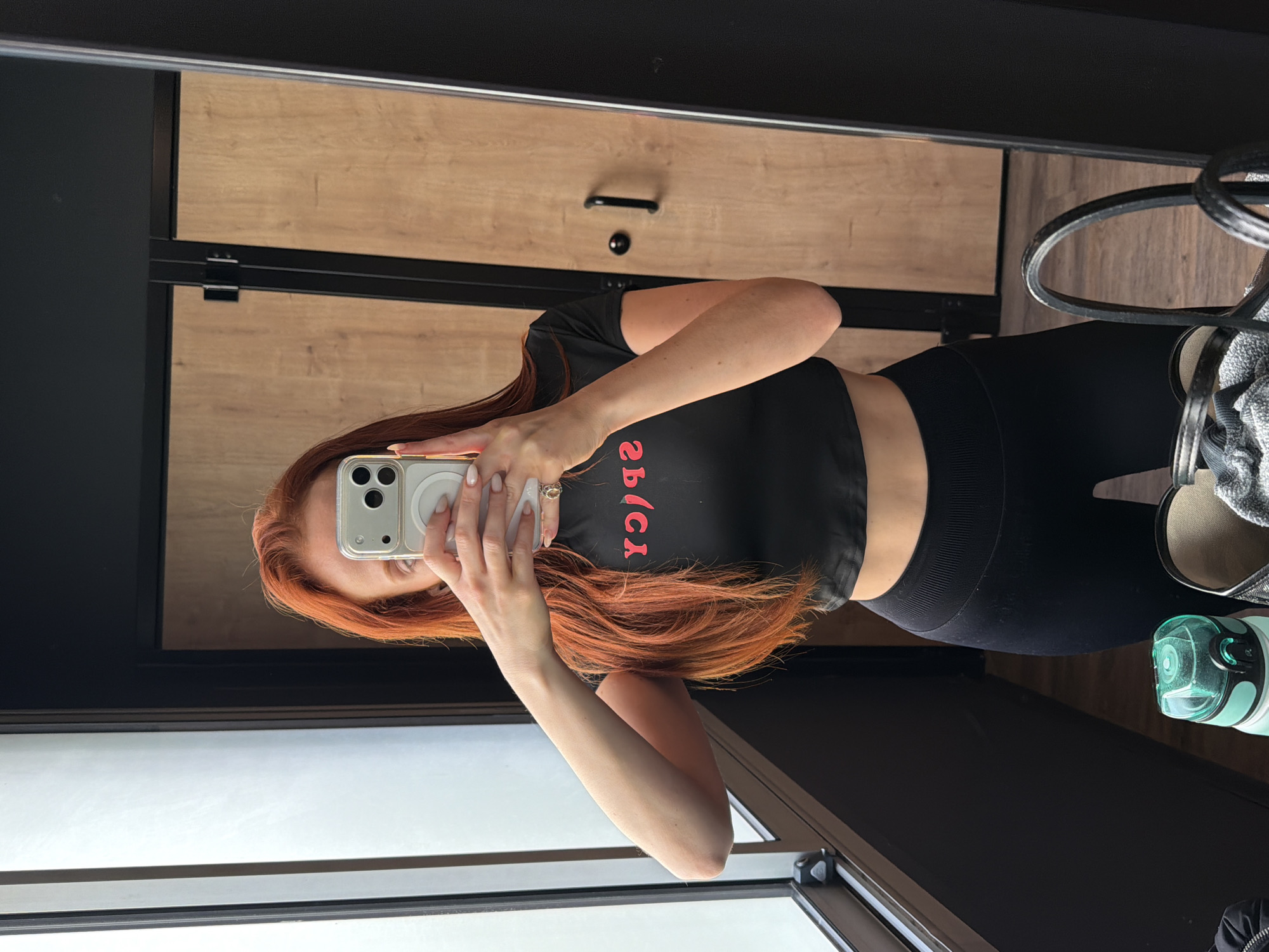 A person with long, red hair is taking a mirror selfie in what appears to be a dressing room or locker room. The person is wearing a black crop top with the word "spicy" written on it in red letters, and black leggings. They are holding a white smartphone up to their face, obscuring their features. The background includes wooden cabinets, a black metal frame, a window, a chair, and a water bottle. There is no indication the photo was taken in Paris, France.