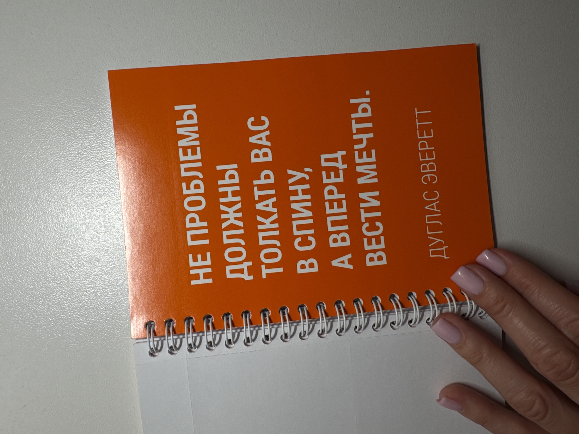 A color photograph shows a spiral-bound notepad lying on a white surface. A hand with pale pink nail polish on each nail is positioned on the right side of the notepad. The notepad has an orange cover, with text printed in white letters arranged in a vertical orientation. The text reads: "НЕ ПРОБЛЕМЫ ДОЛЖНЫ ТОЛКАТЬ ВАС В СПИНУ, А ВПЕРЕД ВЕСТИ МЕЧТЫ. ДУГЛАС ЭВЕРЕТТ". No details suggest this image was taken in the city of Sochi, Russia.