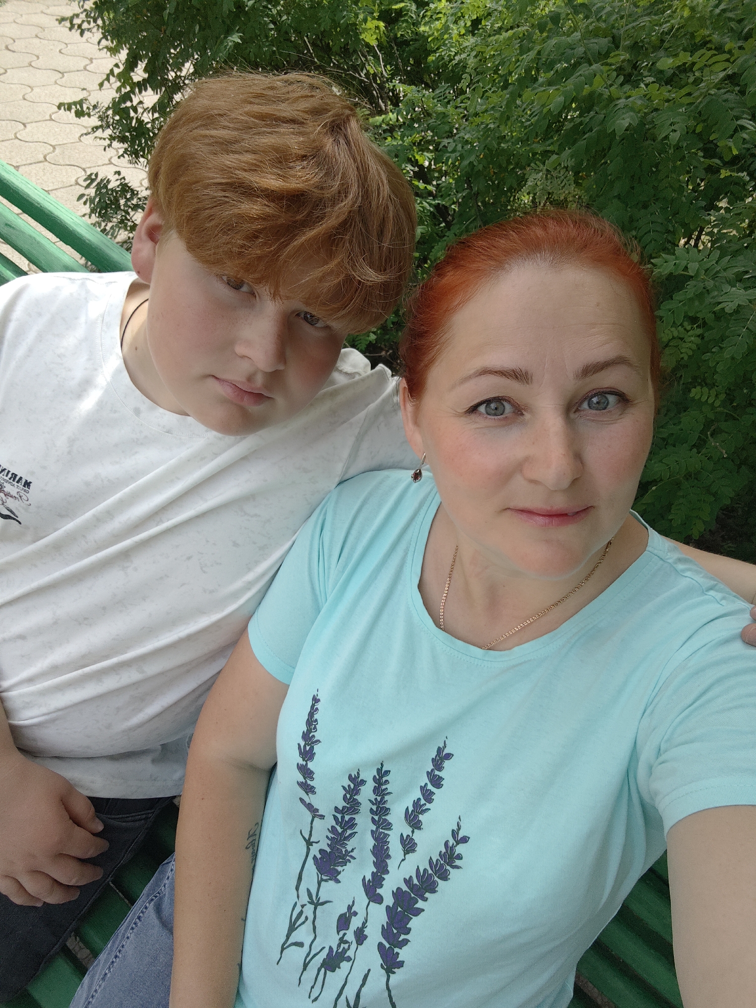 A woman and a boy are sitting together on a green bench outdoors. The woman has red hair pulled back, blue eyes, and is wearing a light blue t-shirt with a plant design on it and a gold necklace. The boy has red hair, and is wearing a white t-shirt. The background includes greenery and a paved area. The location is likely in the city of Chaikovsky, Russia.