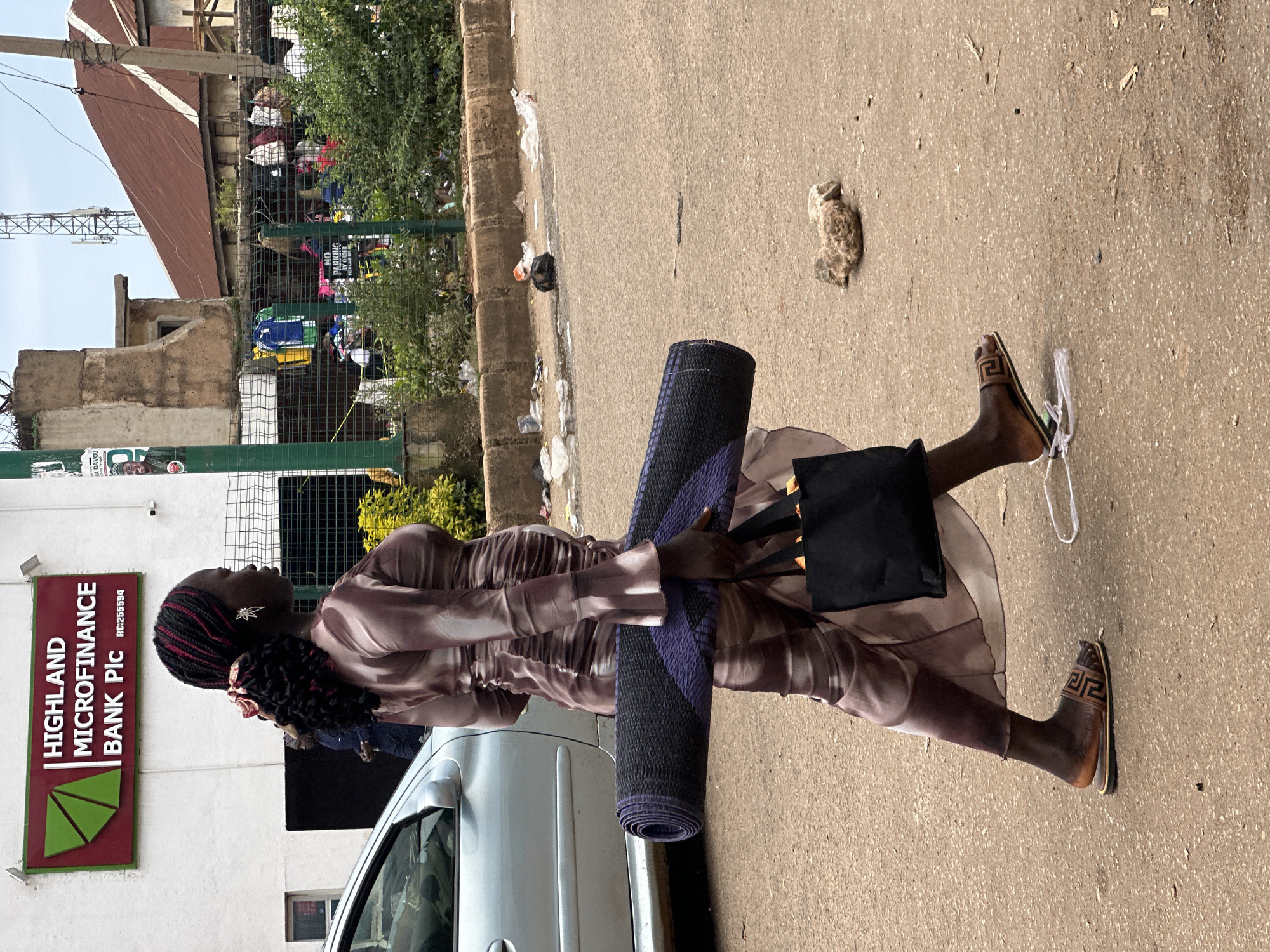 In Bukuru, Nigeria, a woman walks on a dusty road, carrying a rolled-up mat and a black tote bag. She is wearing a long, flowing dress with a marbled pattern and brown sandals. Her hair is styled in braids with red accents, and she wears a star-shaped earring. To her left, a silver car is parked. Behind her, a sign for "HIGHLAND MICROFINANCE BANK Plc" is visible on a building. The environment includes a wire fence with various items hanging on it, overgrown green foliage, and scattered debris on the roadside, including plastic bags and rocks. A telecommunications tower is visible in the background.