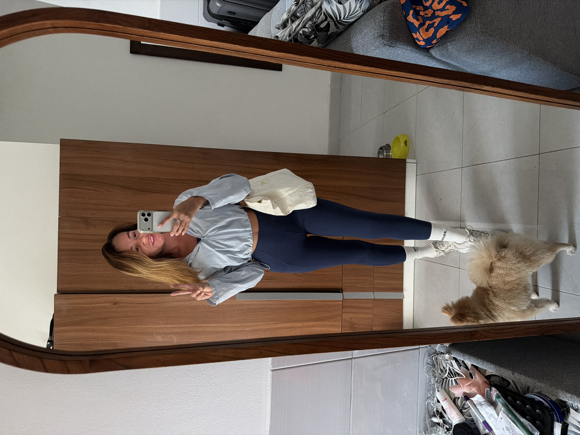 A woman is taking a mirror selfie in a room. She is wearing a light blue cropped zip-up jacket, navy blue leggings, and white socks with white sneakers. She is holding a white tote bag with a palm tree design in her left hand and is making a peace sign with her right hand. She is also wearing a ring on her right ring finger. A small, fluffy tan dog is standing on the tiled floor in front of her. In the reflection, a wooden wardrobe is visible behind the woman. To the right of the woman, a gray sofa with a patterned cushion is visible. Further to the right, a suitcase is leaning against the wall. There is a yellow pet bowl on the floor in front of the wardrobe. The room appears to be a domestic interior.
