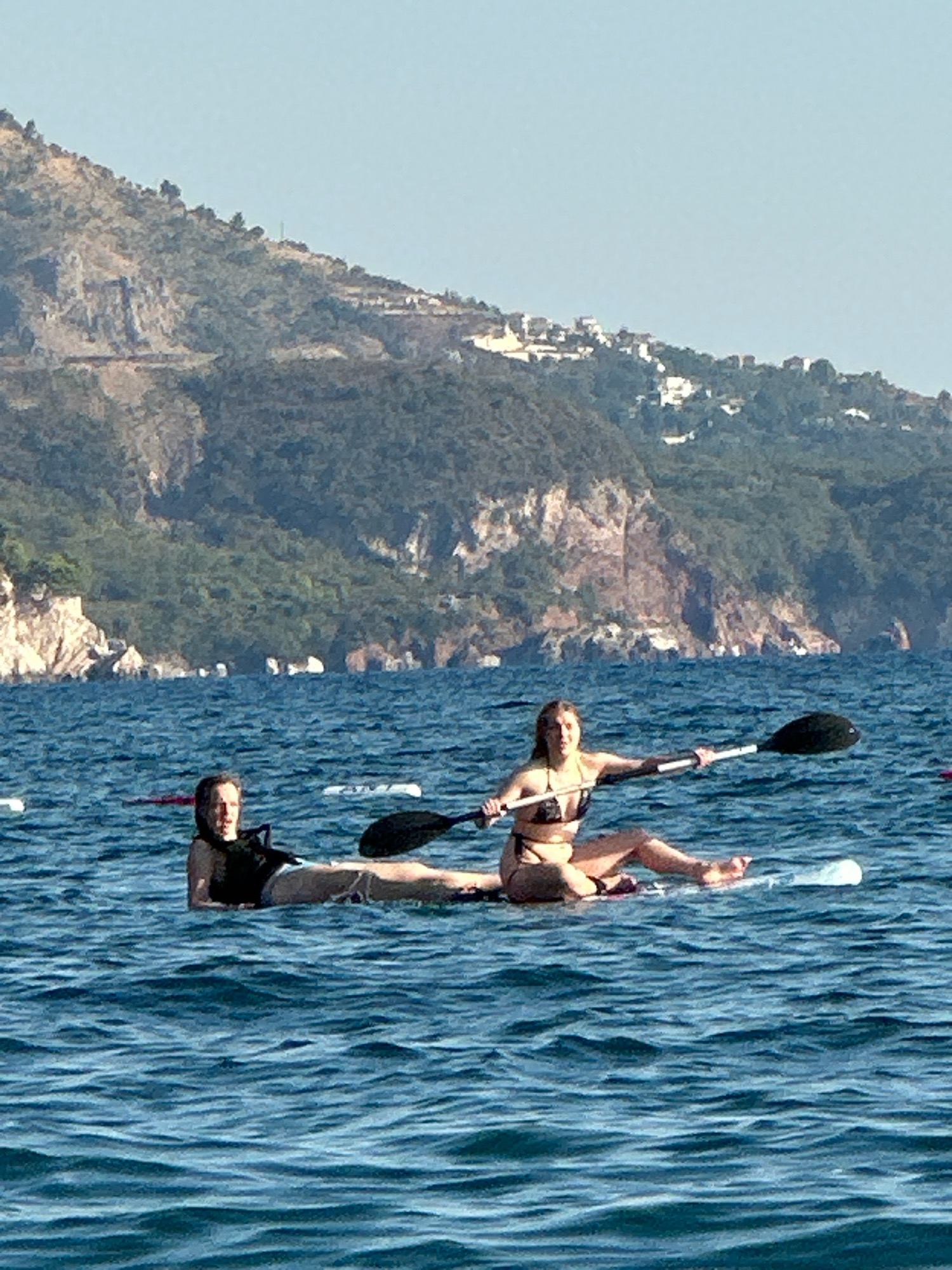 Two individuals are paddleboarding on a body of water with a mountainous coastline in the background. The individual on the left is lying down on the paddleboard, wearing a black life vest over a dark top and shorts. The individual on the right is sitting cross-legged on their paddleboard, wearing a patterned bikini, and holding a paddle. The water is blue and slightly rippled. The coastline is rocky and covered with green vegetation, with some buildings visible on the slopes. The sky is clear and blue. The scene appears to be a recreational activity on a sunny day, likely near Boreti, Montenegro, given the coastal mountain landscape.