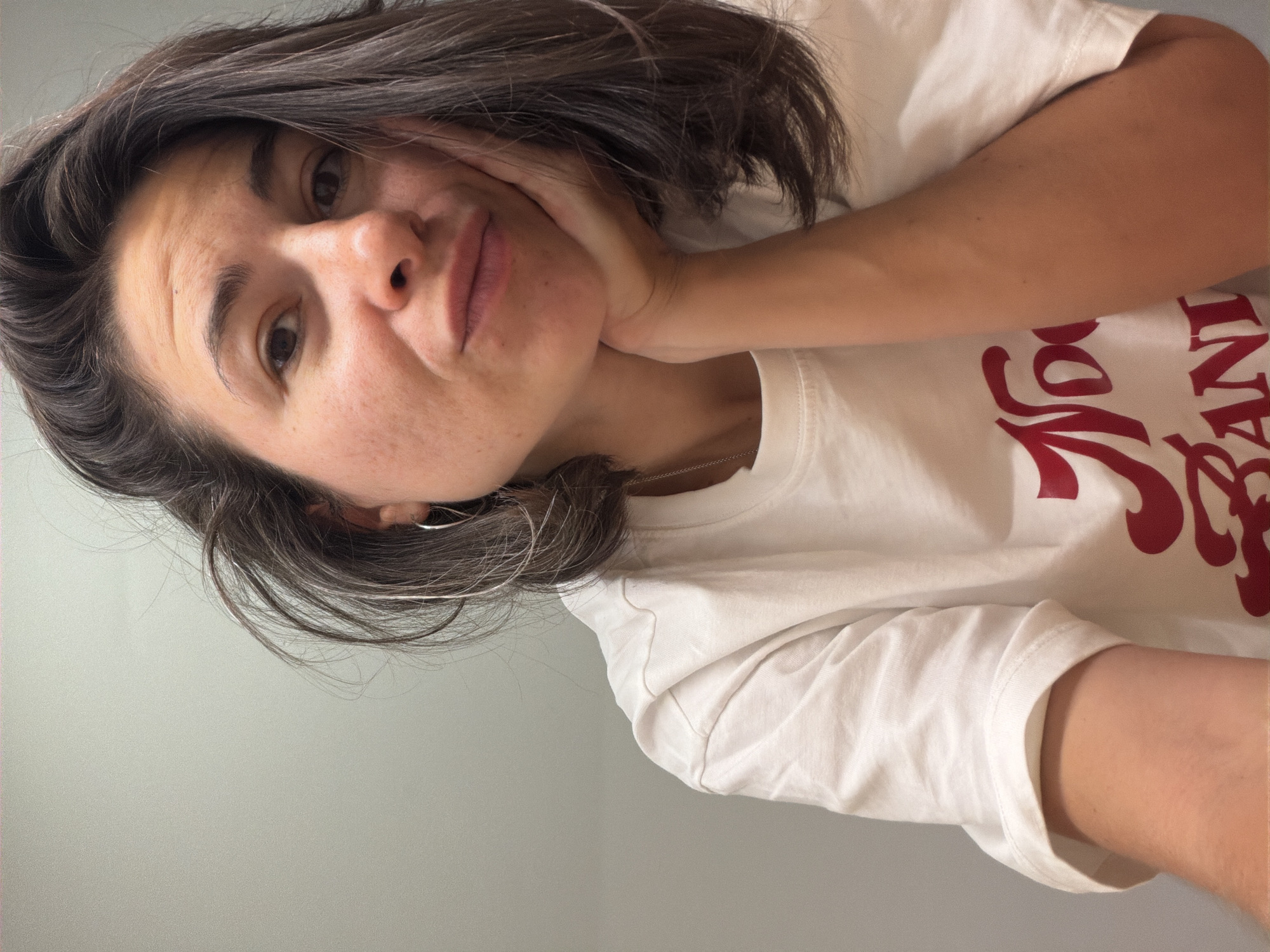 A woman with brown hair and brown eyes is posing for a selfie. She is wearing a white t-shirt with red lettering. The background is a light gray wall. There is no indication of the city of Budva, Montenegro.