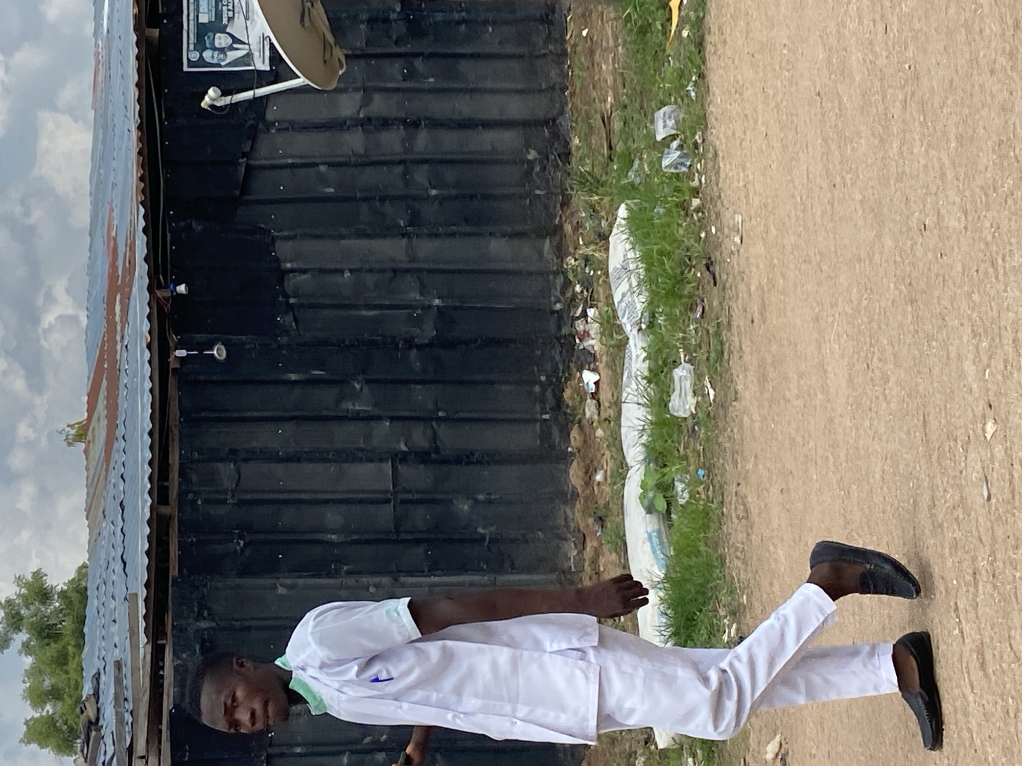 A young Black male, likely a student or health worker, is standing in what appears to be Kuson, Nigeria. He is wearing a white uniform, possibly a lab coat or medical scrubs, with a green collar. The uniform has a blue emblem on the chest. He is also wearing black loafers.
He is standing next to a dark, corrugated metal structure, possibly a building or a shipping container. To the left of the metal structure, a portion of a corrugated metal roof is visible, with some foliage peeking out from behind it. To the right of the metal structure, there is a patch of grass with some scattered white plastic bags and other debris. Beyond the grass is a dirt path.
The man's posture suggests he might be walking or posing for the photograph. He is looking towards the camera with a slight smile. The sky is cloudy, indicating it might be daytime, but the specific time is not clear. The overall scene depicts a humble, possibly rural or semi-urban environment, with signs of both daily life and perhaps some environmental neglect due to the litter. There is a poster visible on the metal structure, but the text is not legible.