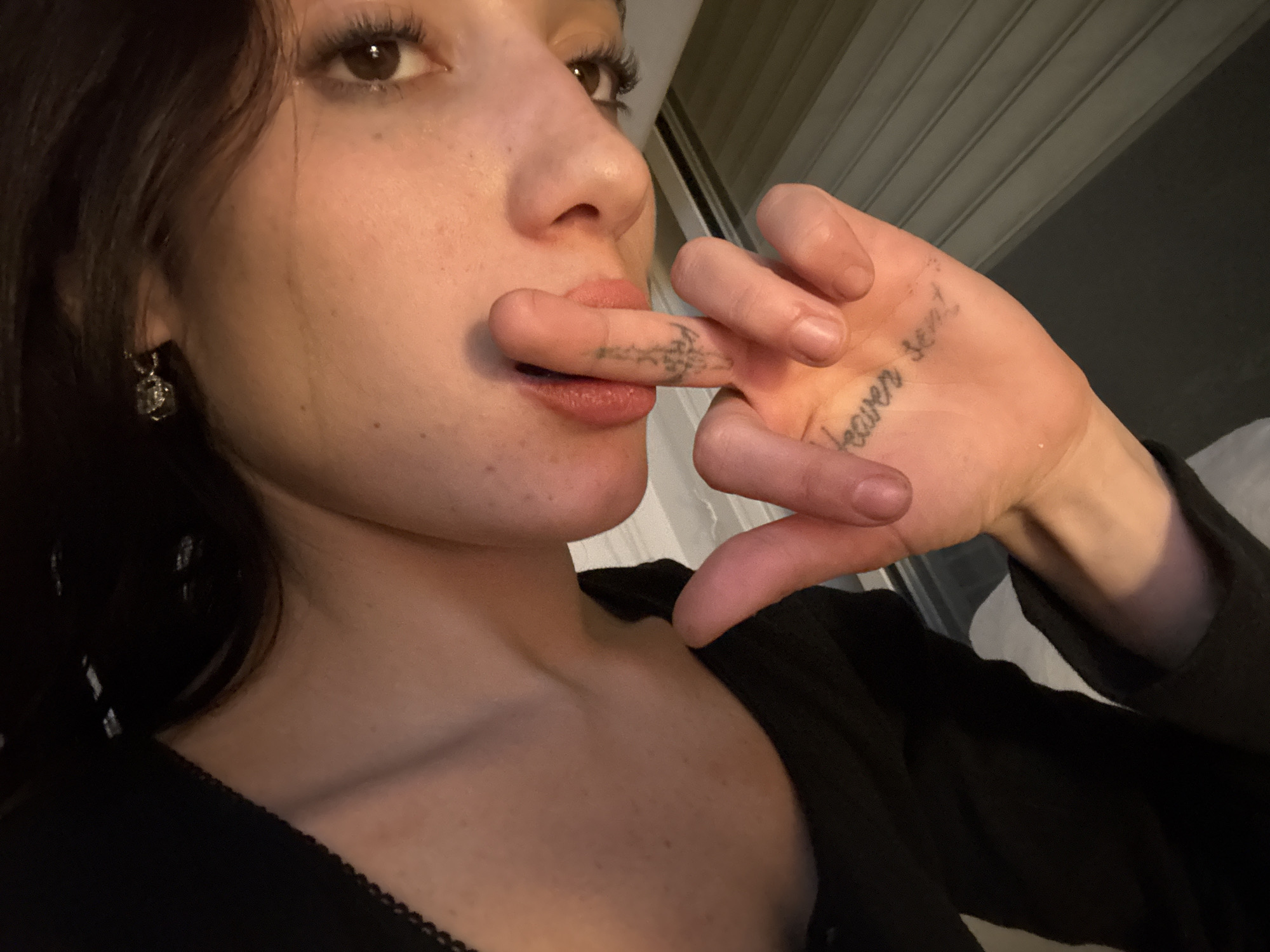 A close-up shot of a woman. She is partially facing the camera with her head turned slightly to the left. Her right hand is raised, with the index finger touching her lower lip. Tattoos are visible on her fingers and the palm of her hand. She is wearing earrings and a black top. The background appears to be an interior space with light-colored walls. The photograph does not contain any information that would allow for identification of its location as Buenos Aires, Argentina.