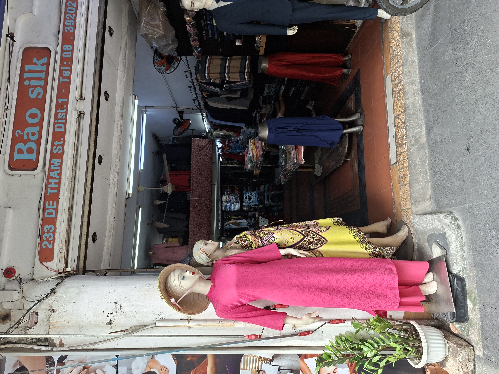 The image shows the storefront of a business named "Bảo silk" located at 233 De Tham St., District 1, with a contact phone number of 08.39202. The storefront displays mannequins dressed in traditional Vietnamese clothing and suits. Two mannequins are prominently positioned at the entrance; one wears a bright pink áo dài and a conical hat, and the other is dressed in a yellow and patterned dress. Inside the store, racks of clothing are visible, along with stacks of fabric. A ceiling fan and fluorescent lighting illuminate the interior. The exterior of the building shows signs of age, with some peeling paint and exposed wiring. The street in front of the store is paved with grey tiles. The overall scene depicts a typical retail environment in a Vietnamese city.