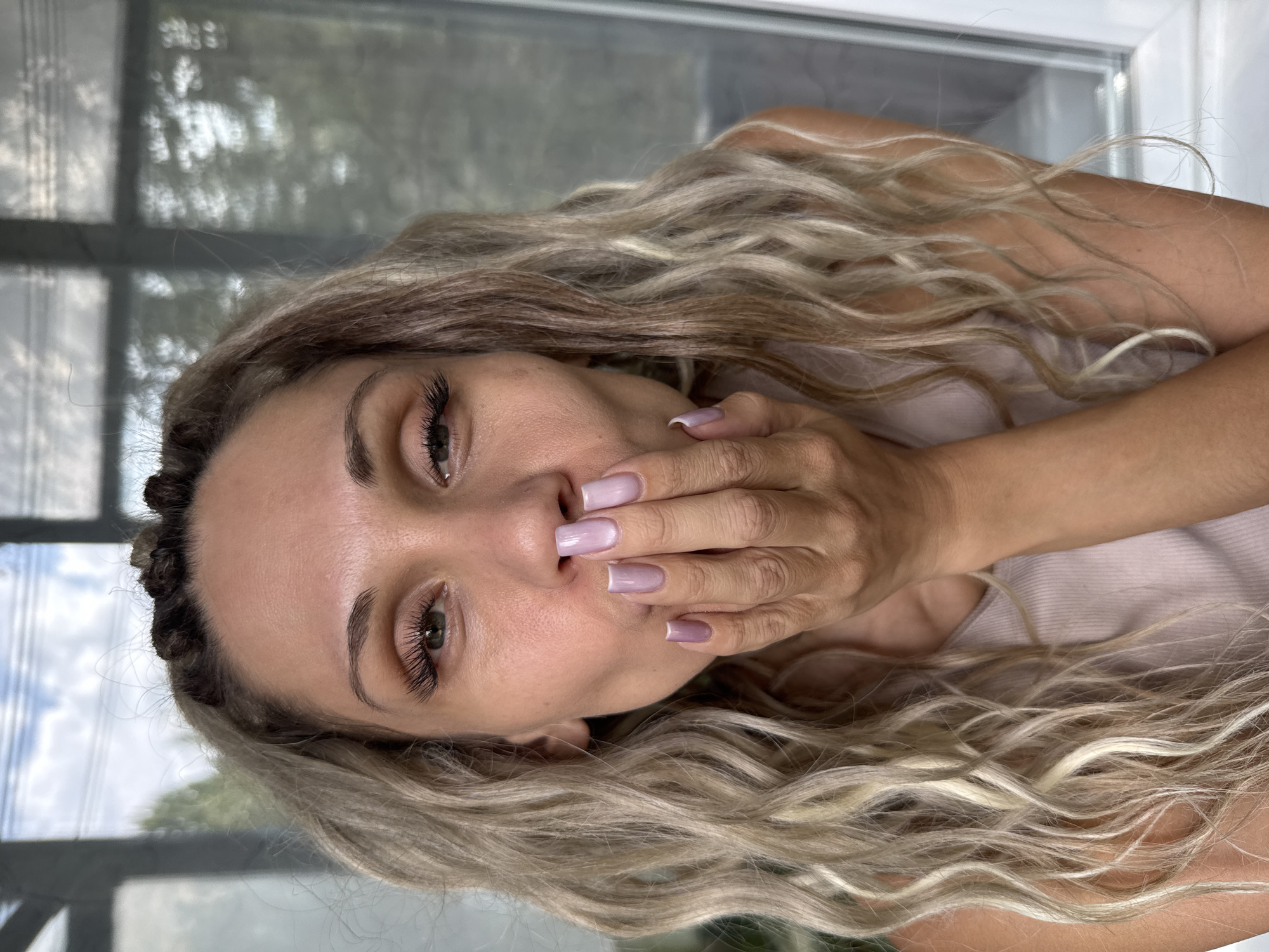 A woman with blonde, wavy hair is pictured from the chest up, facing the camera and puckering her lips as if blowing a kiss. She is wearing a light pink tank top. Her right hand is raised to her mouth, covering her lips and nose, and her fingernails are painted a pale lavender color. Her eyes are looking directly at the camera, and she has long, dark eyelashes. Her hair is pulled back in small braids at the top of her head, with the rest of it falling in waves around her shoulders. The background appears to be an indoor setting with large windows showing a blurred view of trees and a cloudy sky. The woman's skin is lightly tanned. There is no indication of the location being Rusanovo, Russia, or any other specific city.