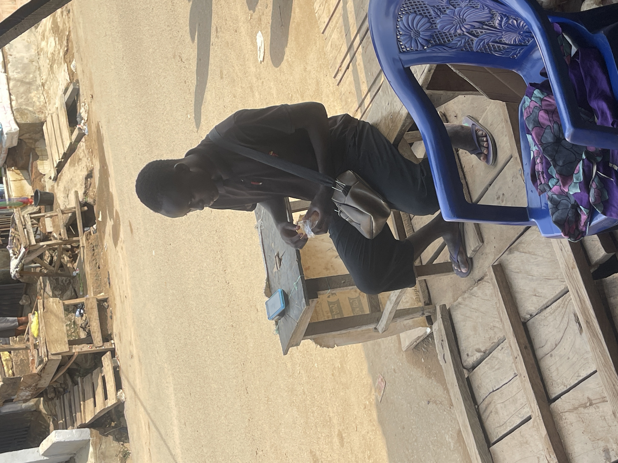 A man is seated at a weathered wooden table on a dusty street in Abuja, Nigeria. He wears a dark t-shirt and dark pants, and a light-colored shoulder bag hangs across his body. He is eating food from a plastic bag. The table appears to be part of a makeshift outdoor shop or stall. A blue plastic chair sits next to the table, with purple and black fabric draped over it. The surrounding area shows buildings and structures made of weathered wood and other materials, typical of a lower-income area or market setting in Abuja. The overall impression is a scene of daily life in an informal urban environment.