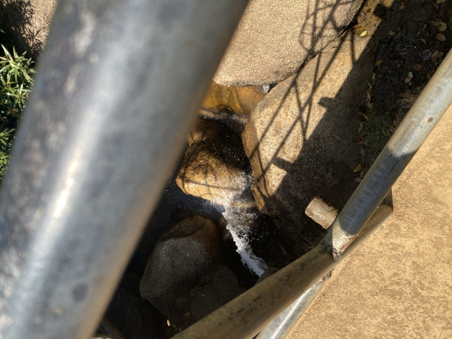 The image displays a downward perspective of a rocky watercourse, viewed through a metal railing. Two weathered, horizontal silver-colored metal bars, part of a railing structure, span the width of the frame, with the upper bar out of focus. A reddish-brown concrete or dirt surface is visible in the lower left beneath the railing. Below the railing, water flows actively over and between several large, rounded rocks, predominantly light brown and grey in color, generating white foam and splashes. Sunlight illuminates portions of the rocks and water, casting distinct grid-patterned shadows onto the rocks and the darker, moist ground beneath them. Sparse green vegetation is discernible in the upper right background. No individuals or identifiable man-made structures beyond the railing are present. The specific location within Kpak, Nigeria, cannot be determined from the visual information provided.