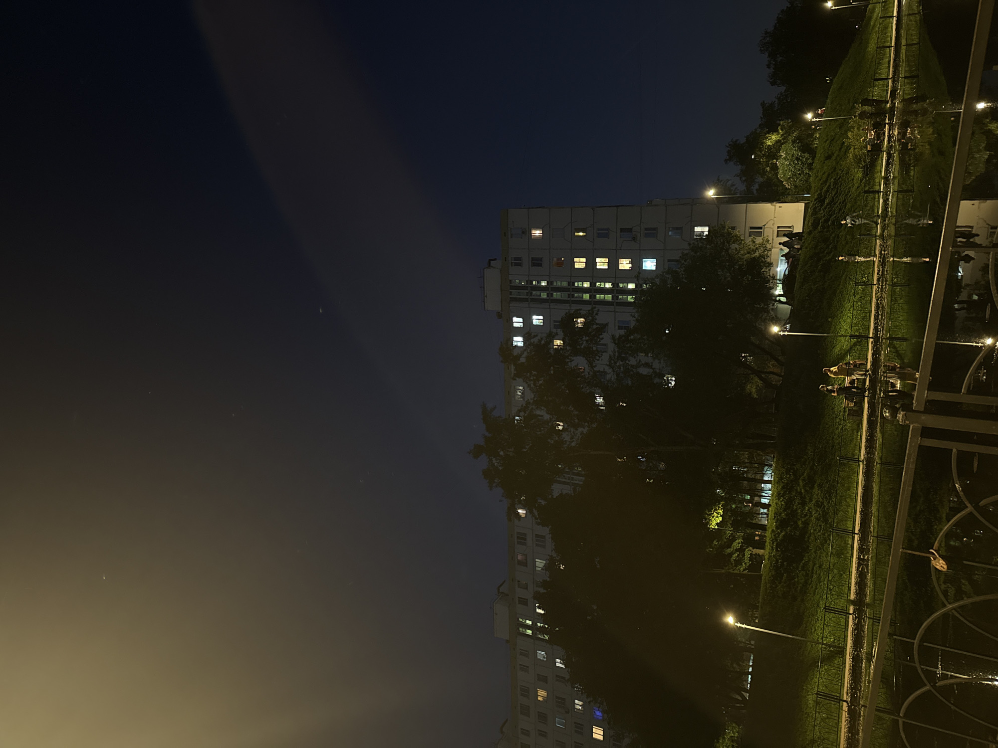 The image depicts a nighttime scene in Moscow, Russia. A tall, apartment building stands illuminated with several lit windows. Trees obscure parts of the building. A park is in the foreground with a fence, and streetlights are visible. A few individuals are walking in the park. The sky transitions from dark blue to a hazy yellow at the bottom.