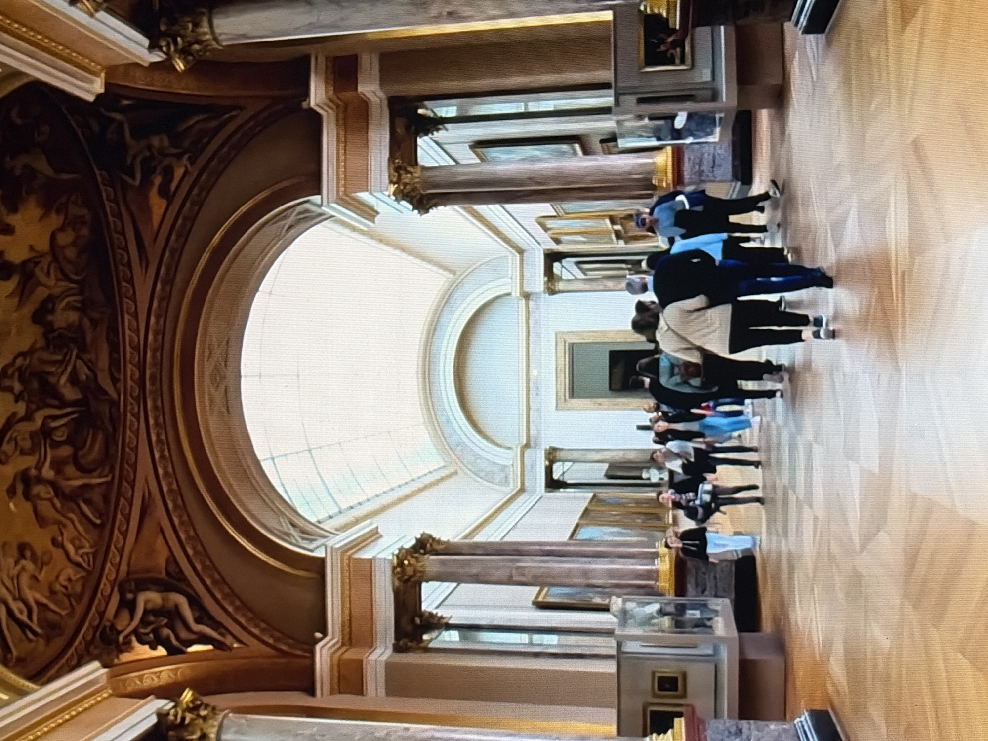 The image shows a long, grand hallway in a museum or art gallery. The hallway features a high, arched ceiling with a skylight that illuminates the space with natural light. The walls are lined with paintings in ornate frames, and large, decorative columns flank the hallway. The floor is made of polished wood with a parquet pattern. Several people are walking down the hallway, appearing to be visitors. They are dressed in casual clothing. The architecture suggests an old-world, classical style, possibly in Europe. The overall impression is one of opulence and cultural enrichment.