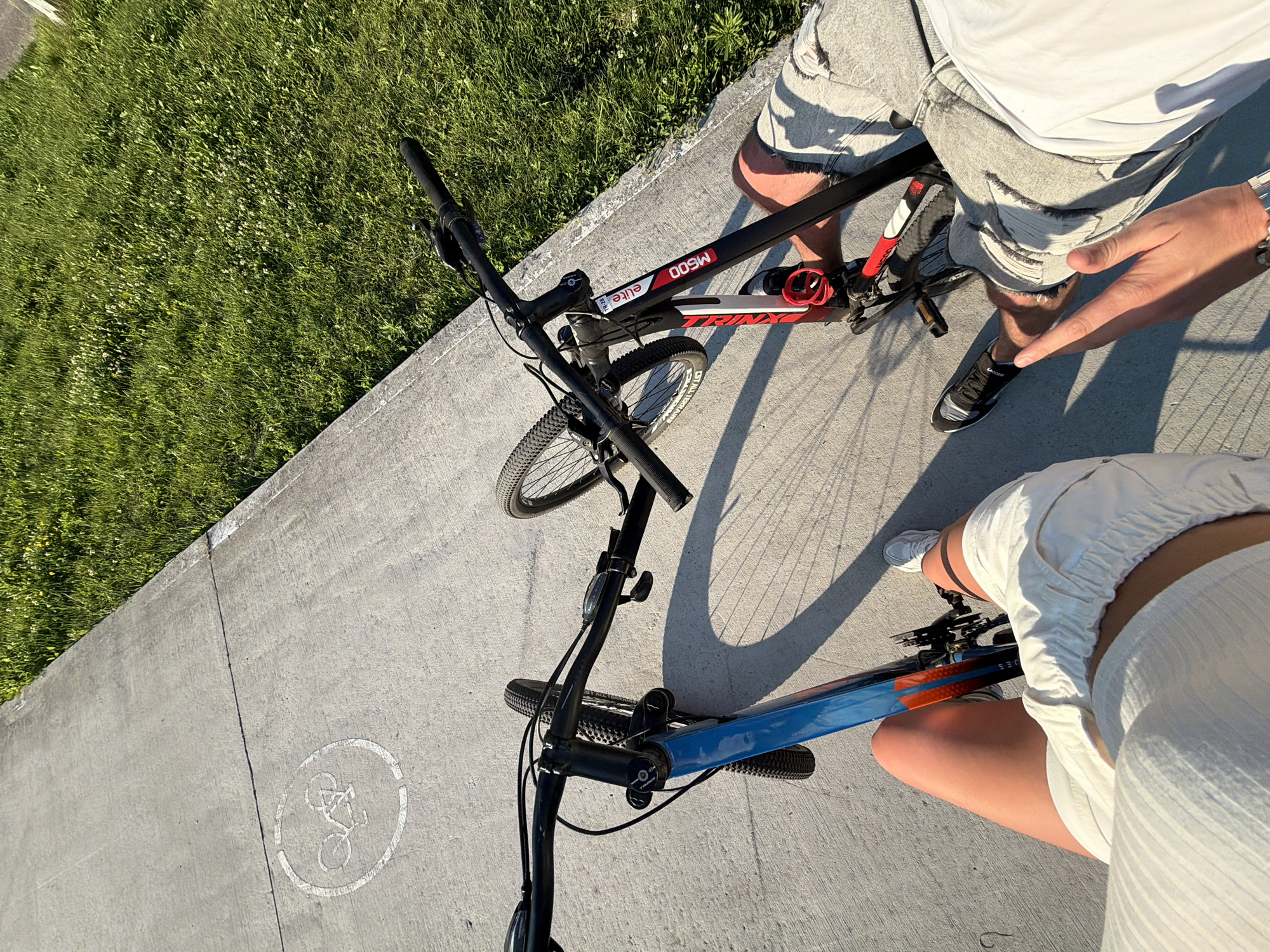 Two individuals are on a paved path, possibly a bike lane, adjacent to a grassy area. One person, wearing white shorts and a white top, is positioned to the left, with their legs and torso visible. Their left hand rests on the handlebars of a blue and orange bicycle. The other person, on the right, is wearing ripped grey shorts and a white shirt, and is partially visible from the waist down. Their right hand is extended towards the viewer, and they are wearing a watch. They are sitting on or near a black and red bicycle. The path has a painted bicycle symbol. The lighting suggests it is daytime, with shadows cast on the path, indicating the sun is relatively low. The environment appears to be an outdoor recreational area, possibly a park or a trail. There is no specific indication of the location being in Kyiv, Ukraine.