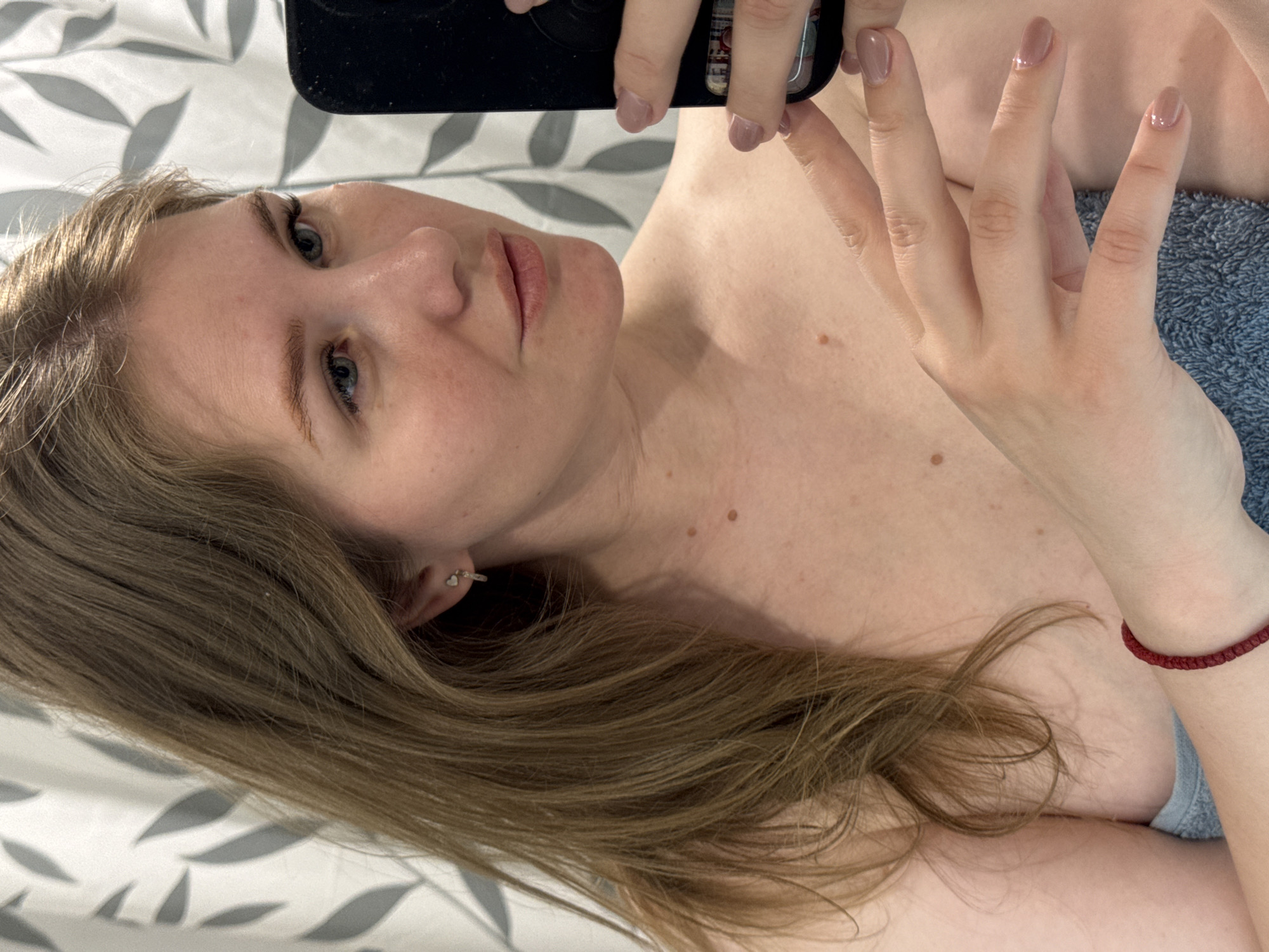 An indoor, overhead-angle selfie captures a young woman lying down, rotated 90 degrees to the left. She has long, light brown hair, blue eyes, and fair skin with several small moles or beauty marks visible on her chest and neck. Her lips are a natural reddish-pink. She wears subtle eye makeup, including mascara. In her left ear, a small, silver-toned earring, possibly a heart or circle stud, is visible.
The woman is holding a black smartphone with her right hand, suggesting she is taking the photograph. Her fingernails are painted a light nude or taupe color. On her right wrist, she wears a red, braided bracelet. A light grey fabric, possibly a top or undergarment, is visible near her shoulder, and a blue, textured fabric, likely a towel, is draped to her right. She appears to be resting on a bed or similar surface, covered by a white sheet with a repeating grey leaf or floral pattern. The expression on her face is neutral and calm.
The scene depicts a quiet, personal moment indoors. There is no visible text in the image. The location context for this media file is Gornyi (Dvurechenskoe S/P), Russia.