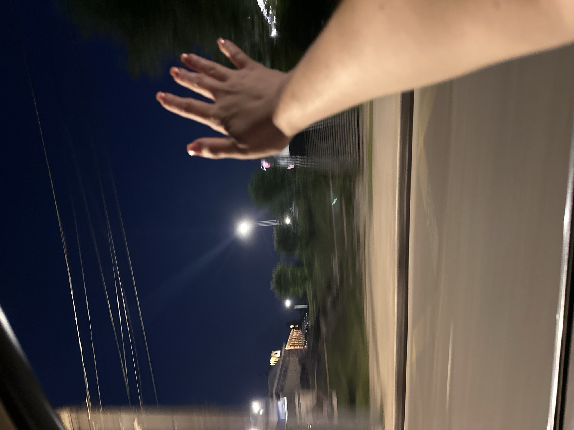 A slightly blurred image shows a hand with orange nail polish partially outside a car window at night. In the background, streetlights illuminate trees and buildings along the road. In the distance, the Pechersky Ascension Monastery in Nizhny Novgorod, Russia is visible.