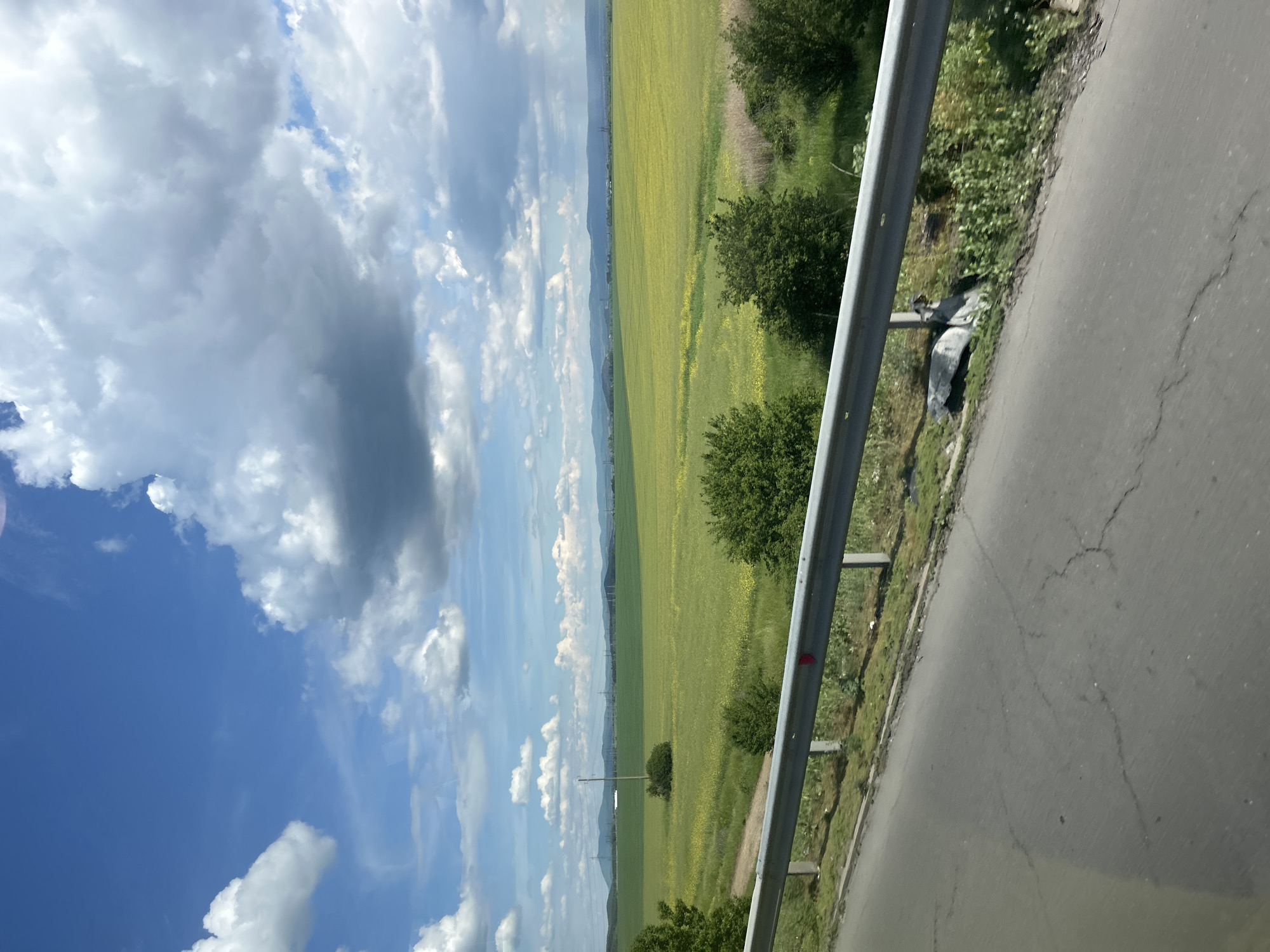 The image shows a landscape with a road, a field, and the sky. The road runs along the right side of the image, separated from the field by a metal guardrail. The field is expansive, green with patches of yellow. The sky occupies the upper left portion of the image and is a mix of blue and white clouds. The exact location of the scene is unknown.