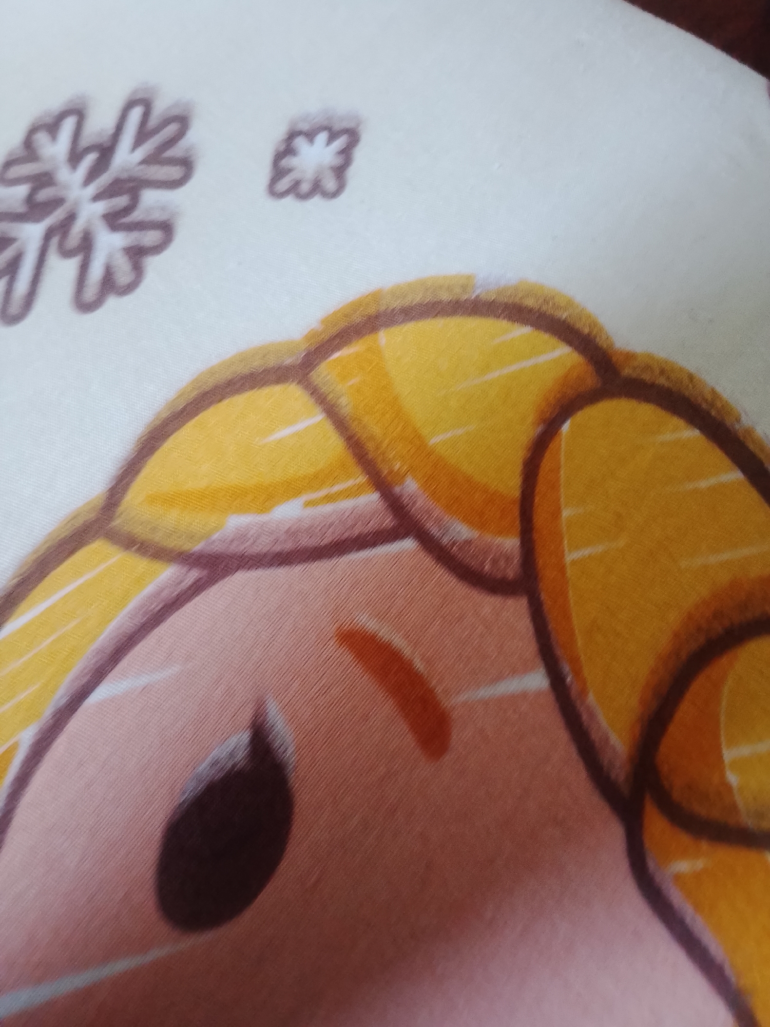 The image is a close-up, angled shot of a white fabric with a printed cartoon character. The character appears to be a young girl with blonde hair, depicted in a stylized manner with large brown eyes. The hair is colored in shades of yellow and orange with brown outlines. There are also small, stylized snowflake designs printed on the fabric in the upper left corner. The fabric itself has a visible texture, suggesting it might be a pillowcase, blanket, or similar textile. The lighting is somewhat dim, creating soft shadows. The image is composed of approximately 80% of the character's face and hair, with the background being the white fabric. No specific location in a city is discernible.