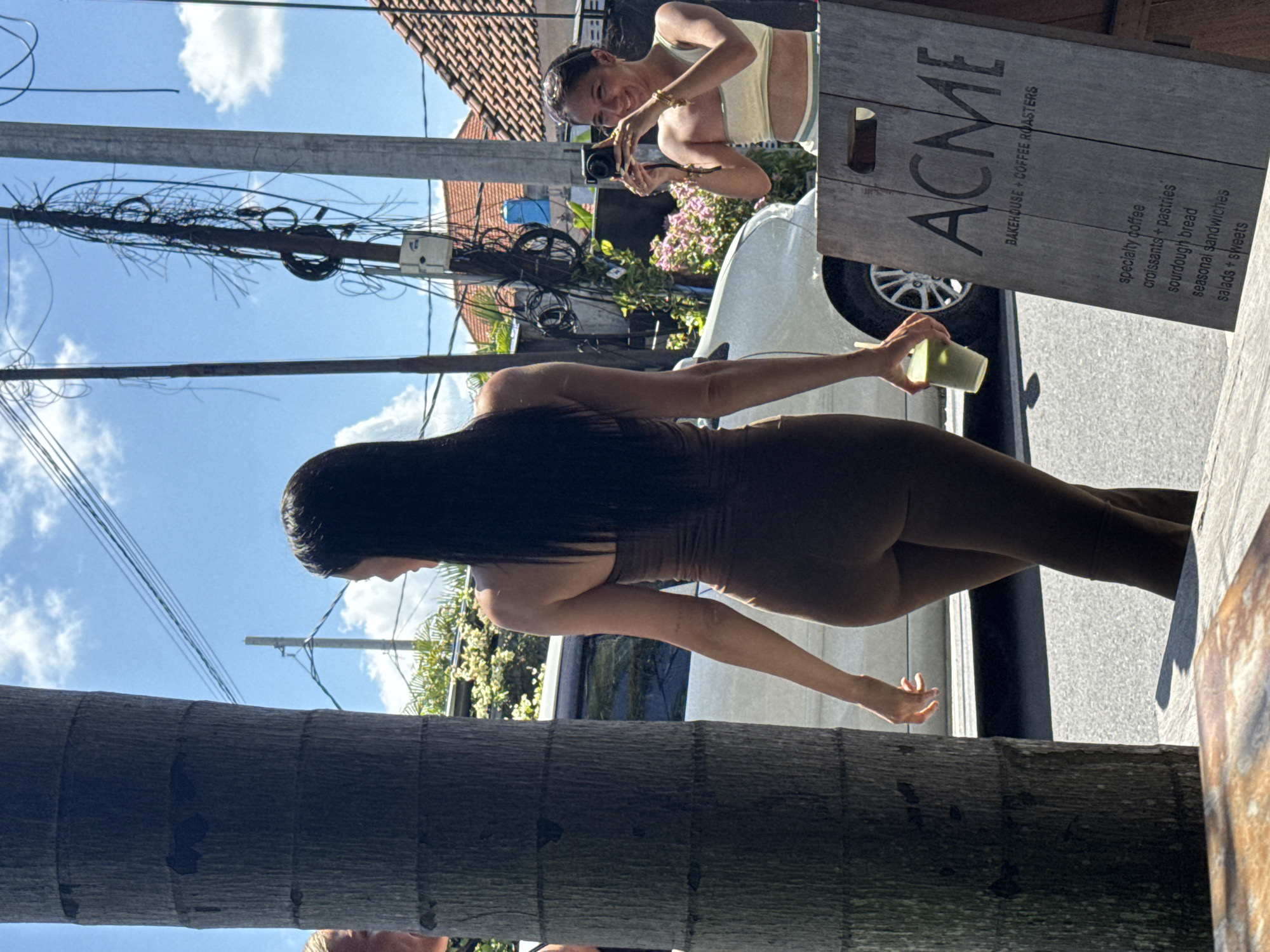 A street scene captured in North Kuta, Indonesia, features two women and a cafe sign. One woman, dressed in a brown jumpsuit, stands near the camera and holds a green drink. Behind her, another woman is reflected in a window, holding a camera and taking a photo. The "ACME Bakehouse + Coffee Roasters" sign is visible beside the reflection, listing specialty coffee, croissants, pastries, sourdough bread, seasonal sandwiches, and salads/sweets. A tree trunk dominates the foreground on the left, while electrical wires and buildings are visible overhead. A white car is parked near the cafe sign.