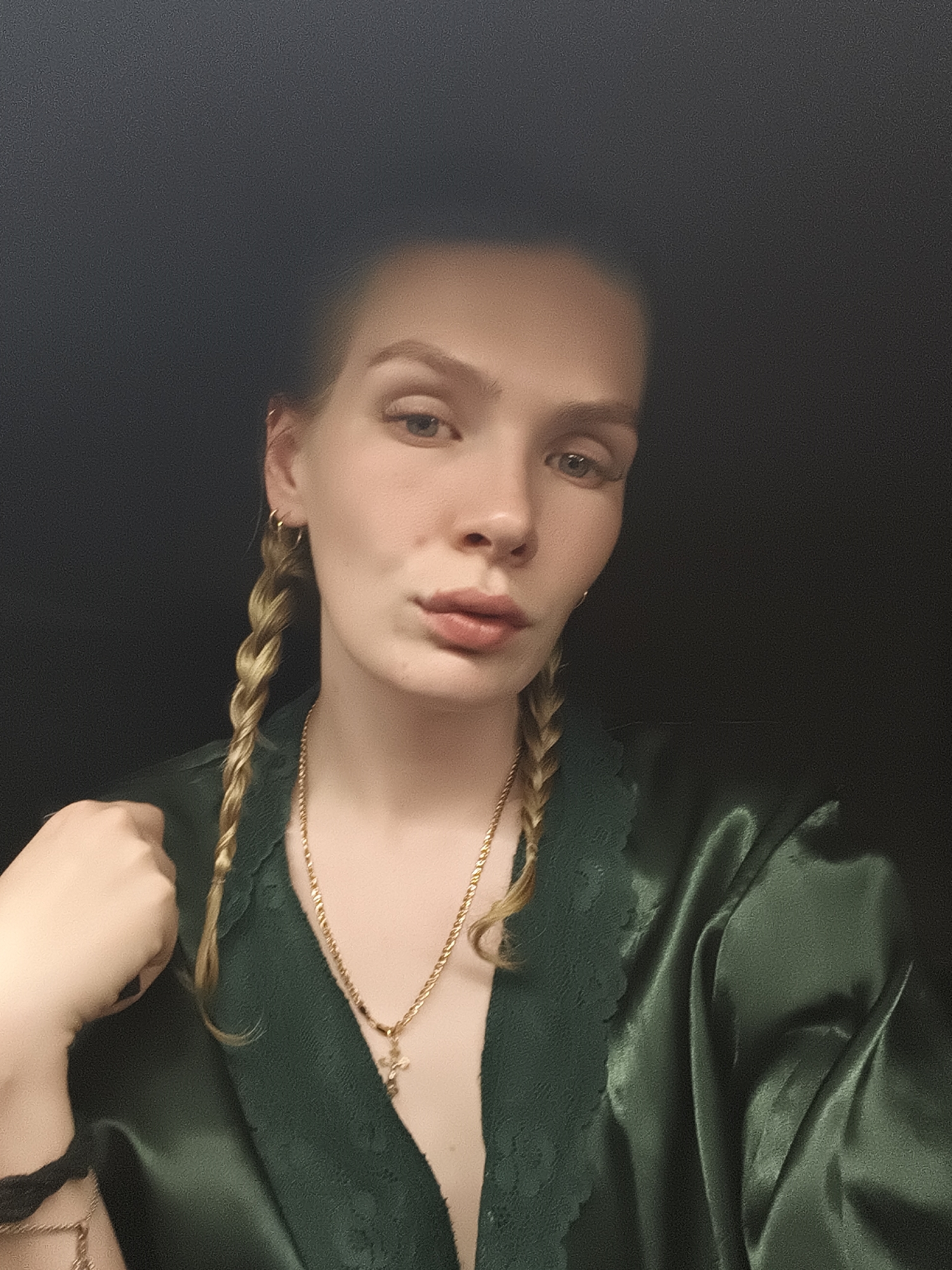 A fair-skinned woman with blonde hair braided into two pigtails is the primary subject of the image. She is wearing a dark green satin garment with lace detailing around the collar, and a gold necklace with a cross pendant. Her left hand is partially visible in the lower left corner of the frame. The background is a uniform dark gray or black. The location is not discernible and cannot be identified as Русский Вожой, Russia based on the available information.