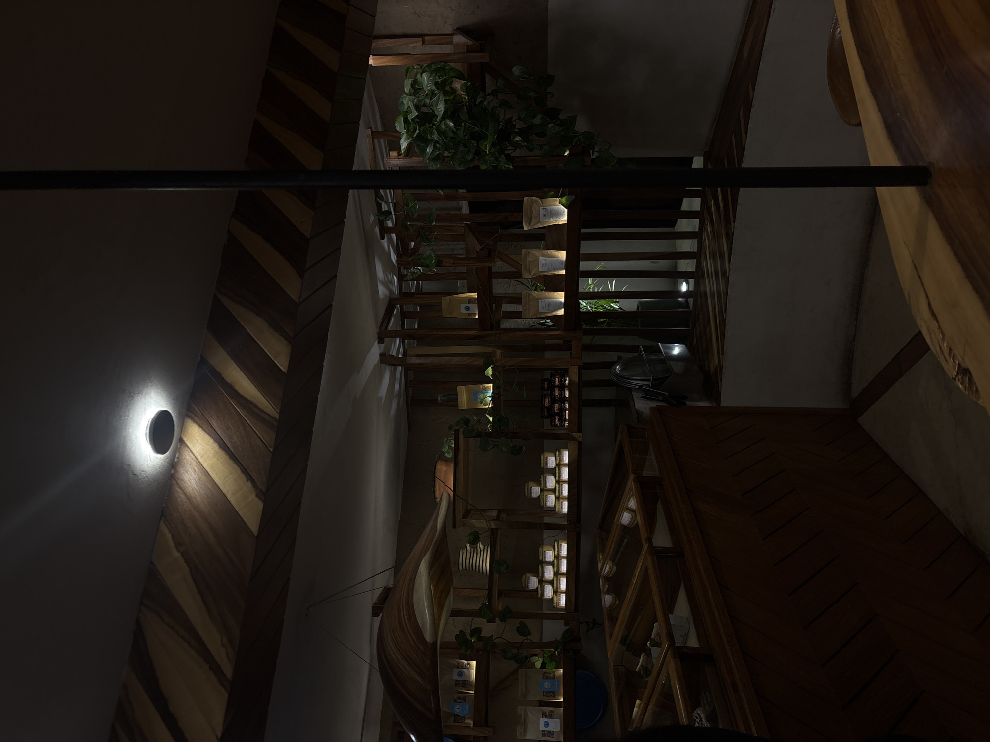 The image shows an interior scene of a café or restaurant in San Andrés Cholula, Mexico. The space features a combination of architectural elements and decorative items. The walls are a mix of painted surfaces and herringbone-patterned wooden panels. A circular wall sconce provides illumination on the left side of the frame.
A series of wooden shelves are filled with various items, including plants, bags of coffee or similar products, and decorative objects. These shelves are arranged in a staircase-like fashion. A canoe or boat-shaped decorative element is also visible. The overall lighting in the space is dim, creating a warm ambiance. A black rod bisects the image horizontally. A part of a wooden table is visible on the right edge of the picture.