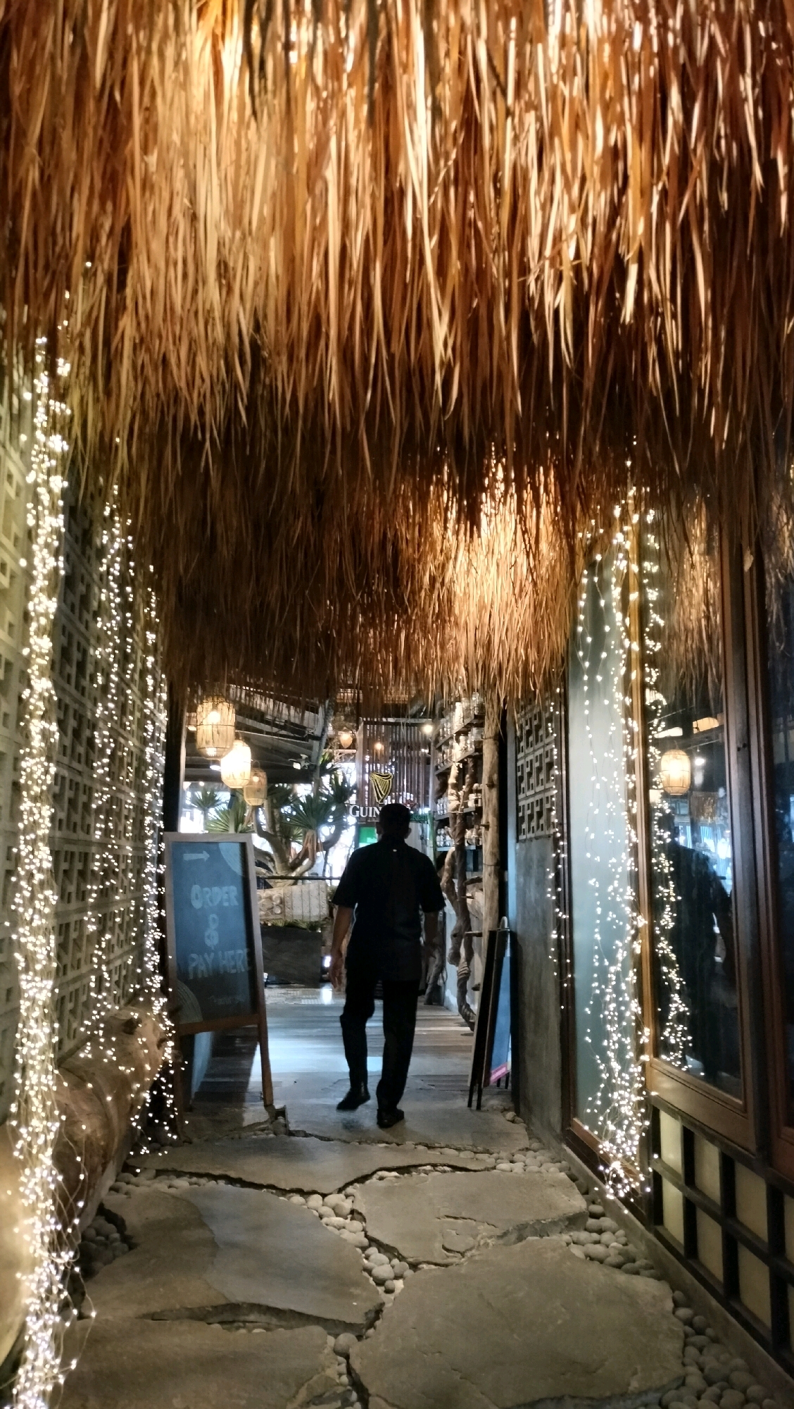 A person walks away from the camera down a pathway in what appears to be a restaurant or bar in Berastagi, Indonesia. The pathway is made of large, flat stones interspersed with small pebbles. The pathway leads through an entryway covered with a thatched roof made of dried grass. String lights are draped along the walls on either side of the pathway, and lanterns hang from the thatched ceiling. A chalkboard sign with "ORDER & PAY HERE" written on it stands to the left of the pathway. The person is wearing a black shirt and black pants. The environment is dimly lit, with the string lights providing most of the illumination.
